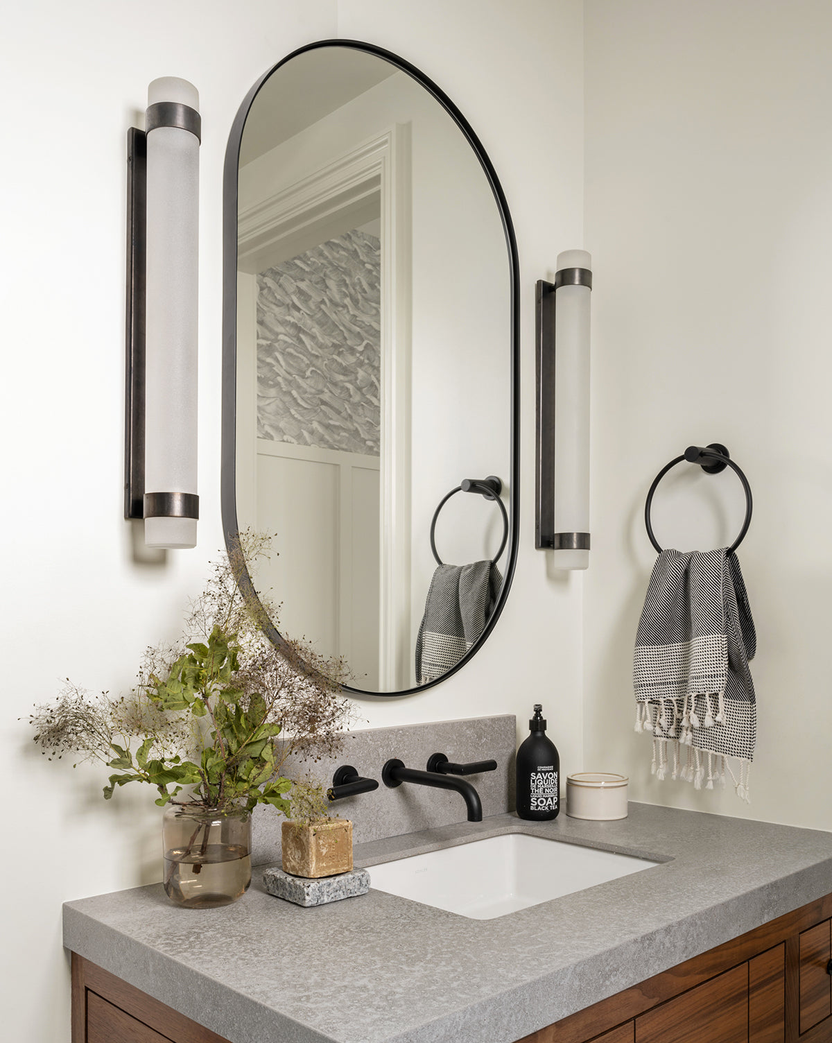 Modern bathroom vanity with the Rye Wall Mirror, vertical wall lights, gray countertop, rectangular sink, stainless steel soap dispenser, black faucet, potted plant, towel ring with gray towels, and decorative accents, by McGee & Co.
