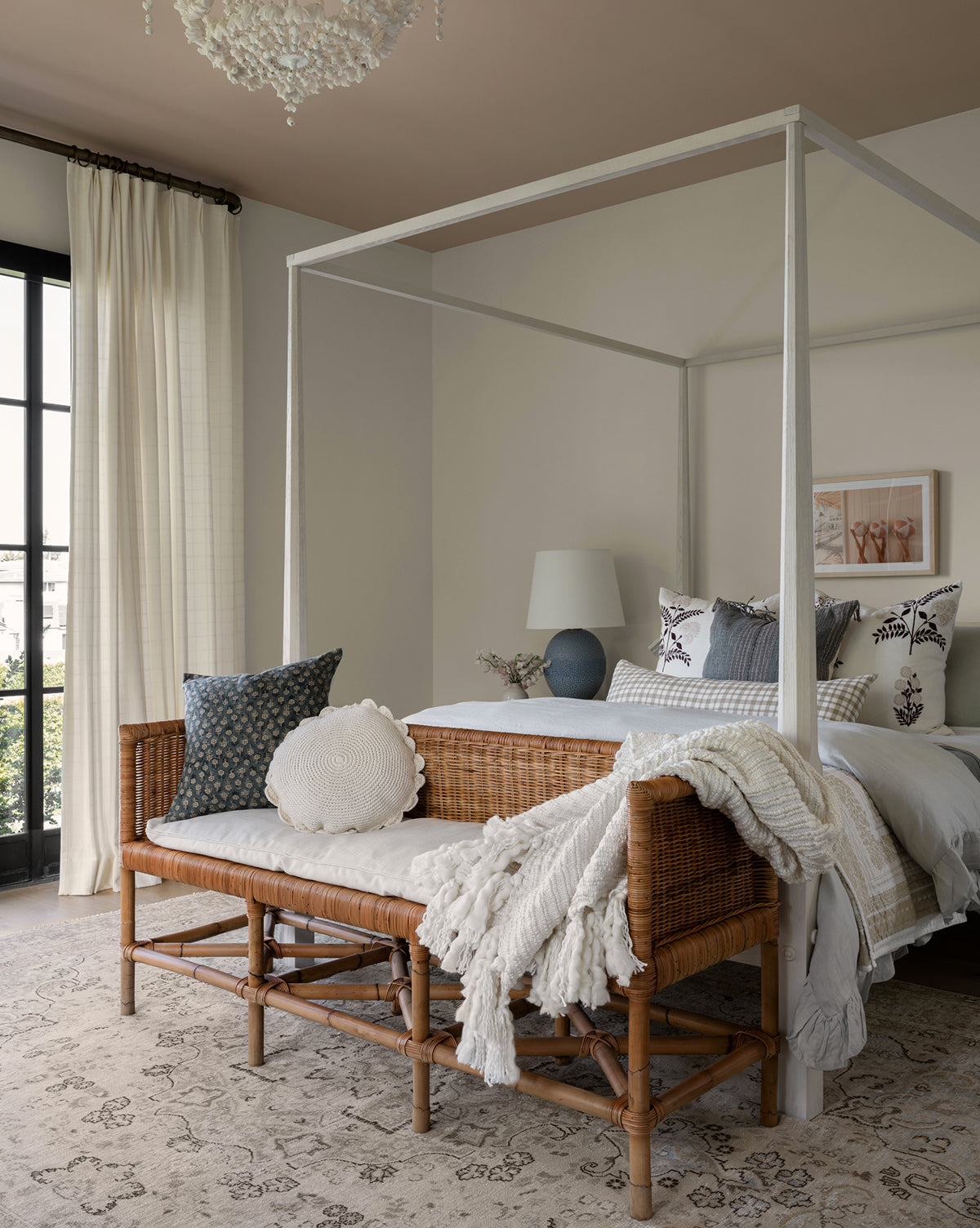 A cozy bedroom with a white canopy bed, performance fabric pillows, the Odelia Bench at the foot, neutral bedding, a soft rug, and a table lamp. Light curtains cover tall windows, and wall art hangs above the bed.