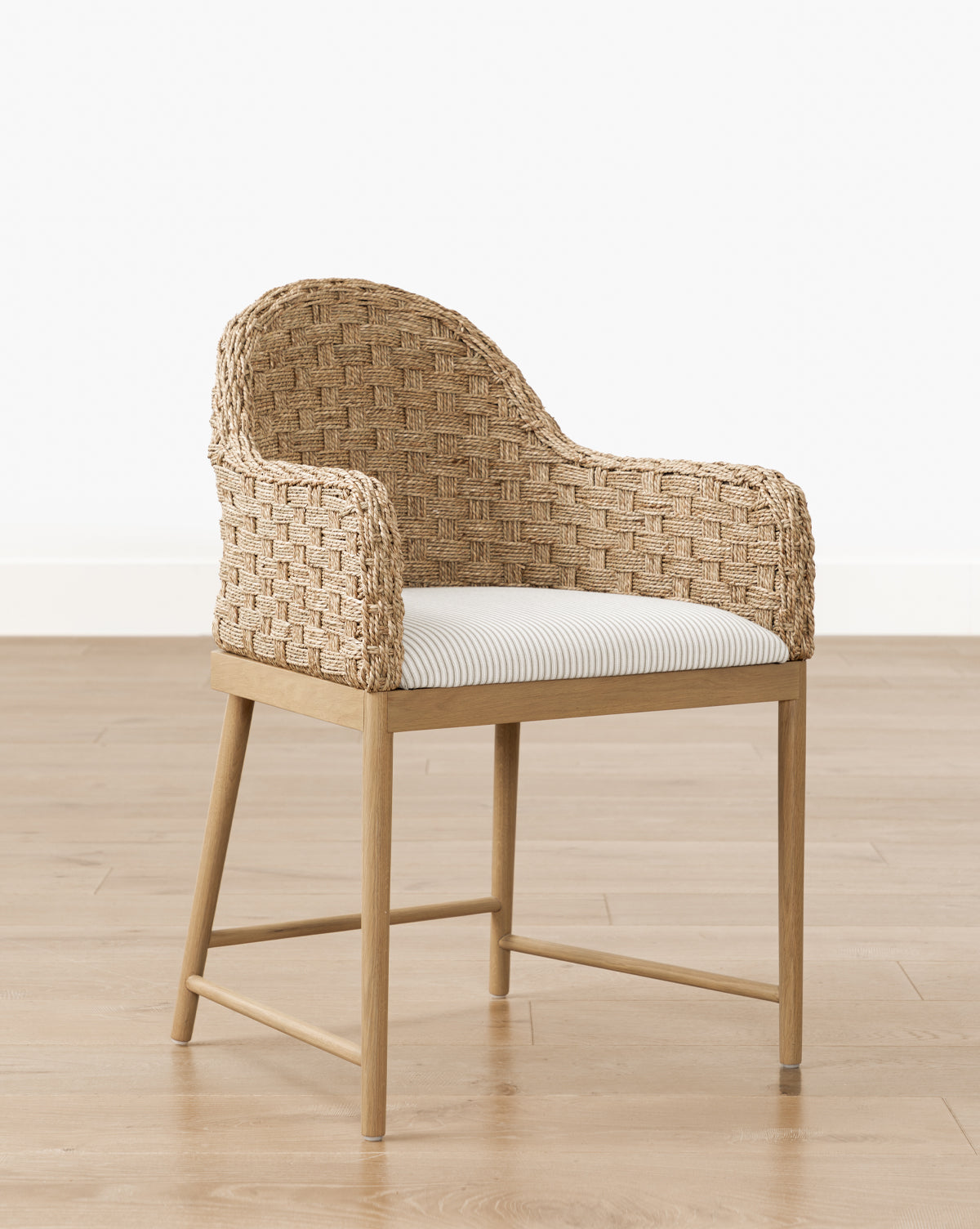 The Molly Arm Chair is a modern woven rattan chair with a curved back and armrests, set on a light wood frame, and includes a white cushion with thin gray stripes. It’s shown on a light wooden floor against a white wall by McGee & Co.