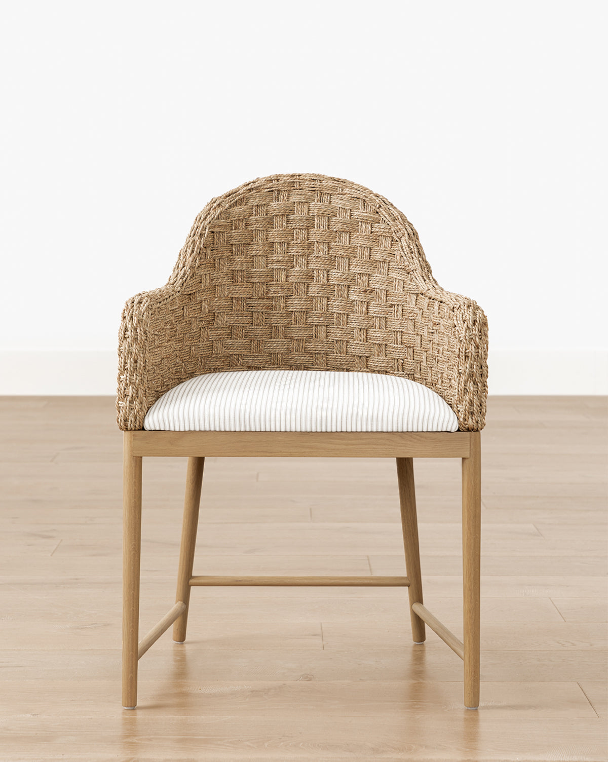 The Makerspalm Molly Arm Chair features woven rattan, a curved back, white striped cushion, and solid oak legs, shown on a light wood floor against a plain white wall. McGee & Co.