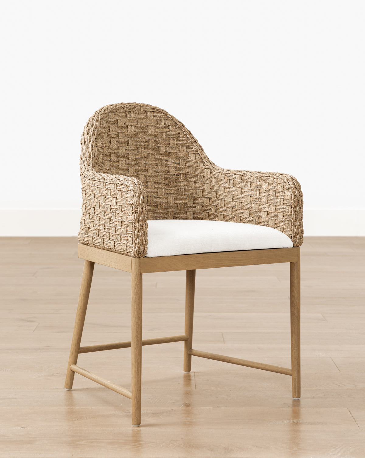 The Molly Arm Chair features a modern woven rattan design with curved back and arms, a light wood frame, and a white cushioned seat, set on a light wood floor against a plain white wall by McGee & Co.