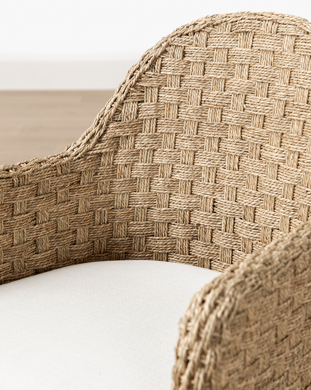 Close-up of the Molly Arm Chair’s woven rattan armrest and backrest in a basketweave pattern, paired with a light cushion. This modern chair rests on a wooden floor with a softly blurred background for emphasis, by McGee & Co.