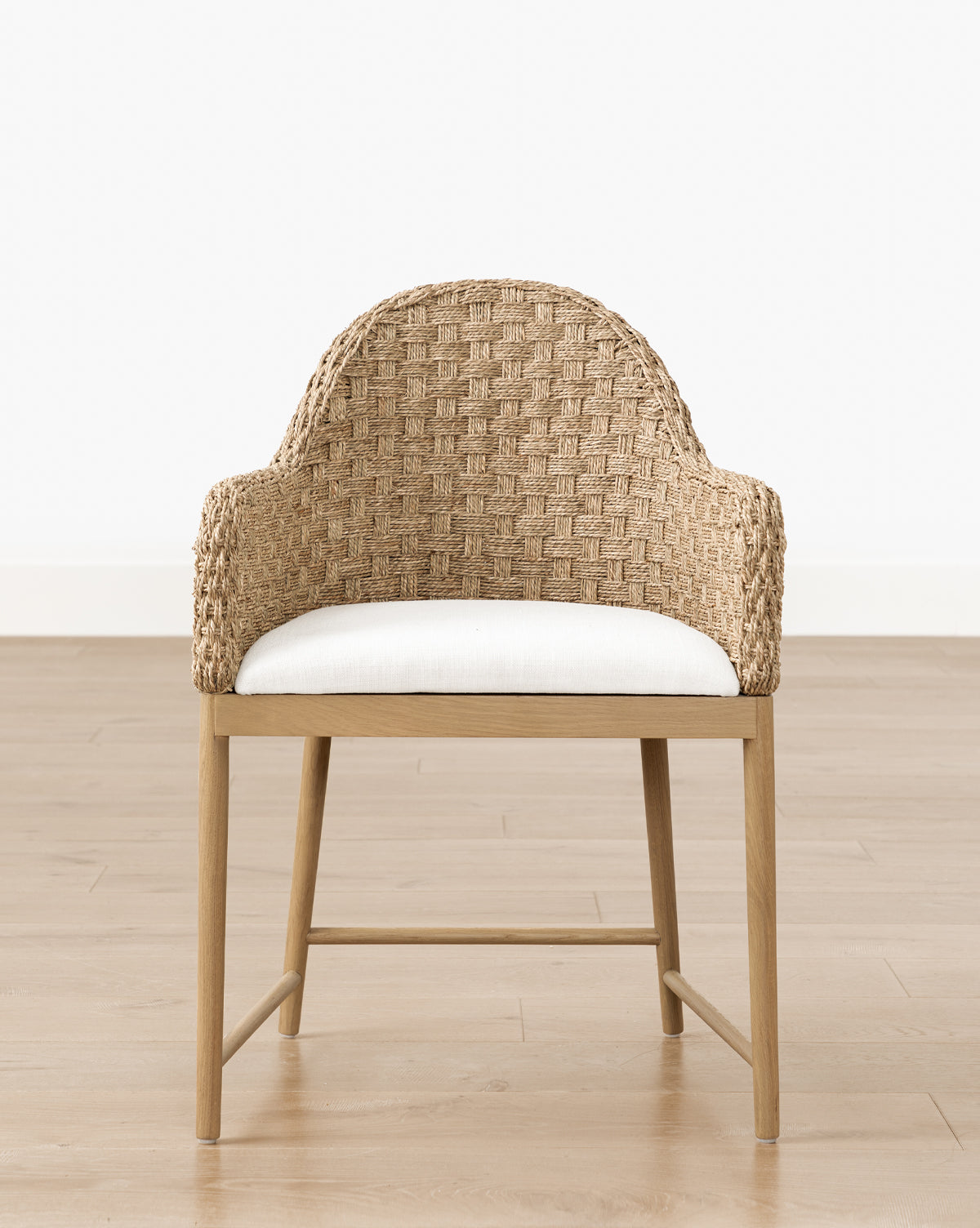 The Molly Arm Chair features a woven rattan design, curved backrest, wooden legs, and a white cushioned seat, shown on a light wood floor with a plain white wall in the background by McGee & Co.