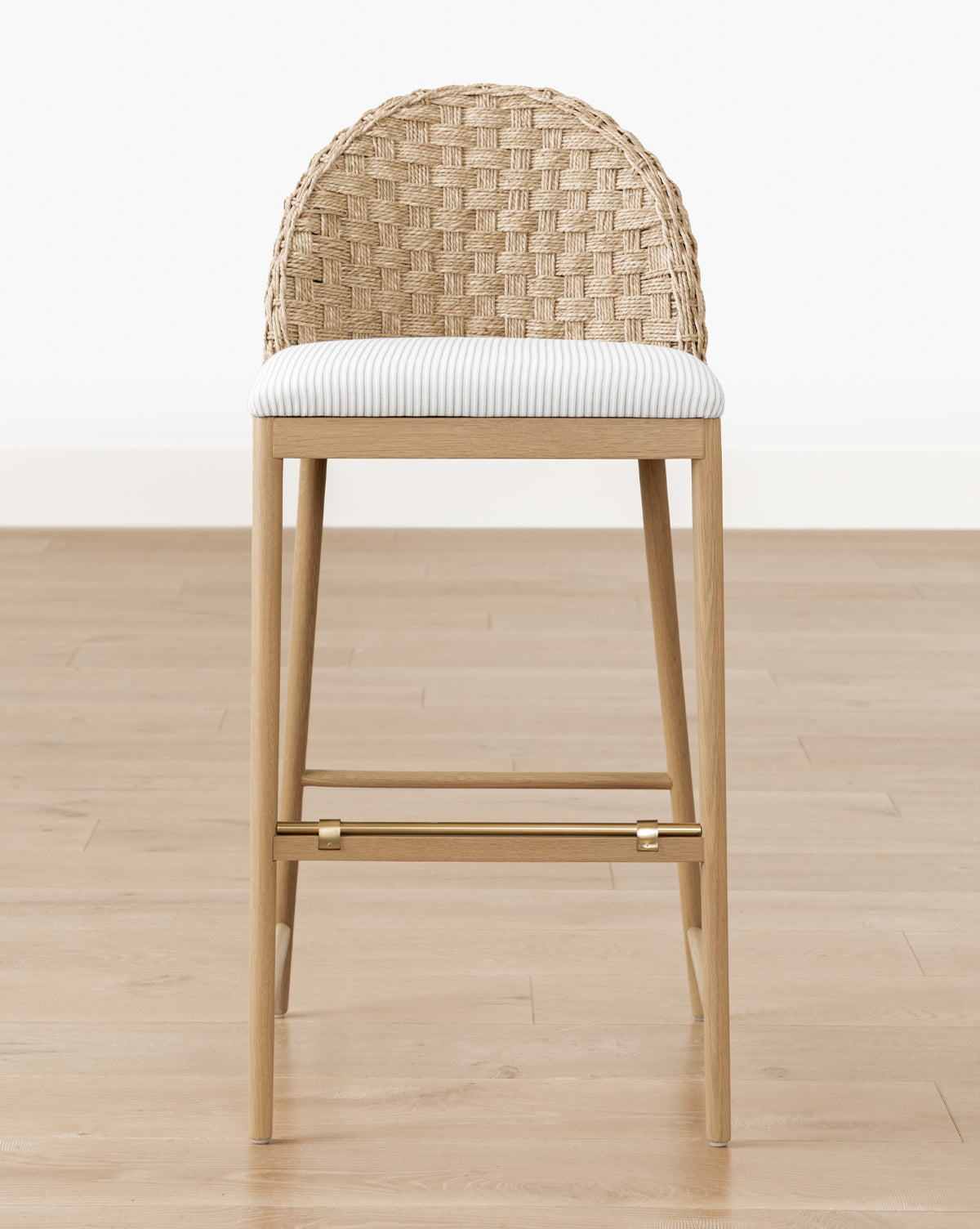 The Molly Stool features solid oak legs, a cushioned light striped seat, and a woven rattan curved backrest. Perfect against a white wall and wooden floor, it adds warmth and style to any space by McGee & Co.