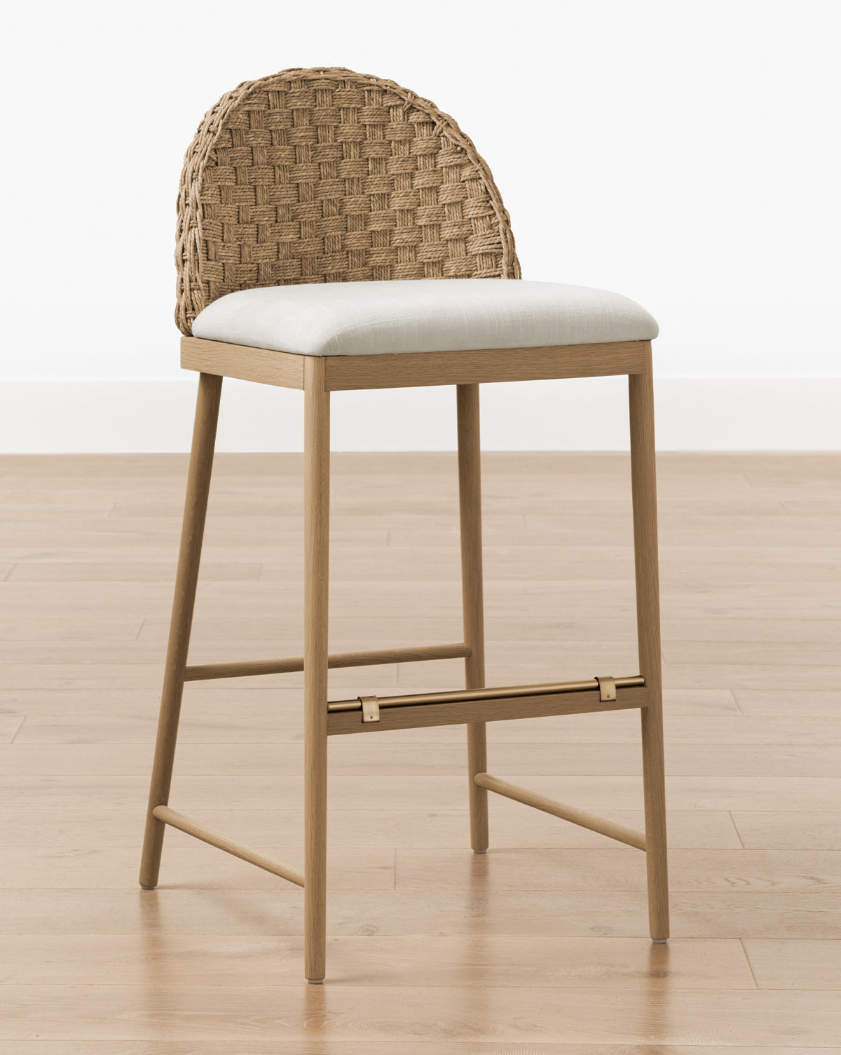 The Molly Stool features a solid oak frame with four legs, a woven rattan curved backrest, a light beige cushioned seat, and a metal footrest, by McGee & Co. Shown on a light wood floor against a plain white wall, by McGee & Co.