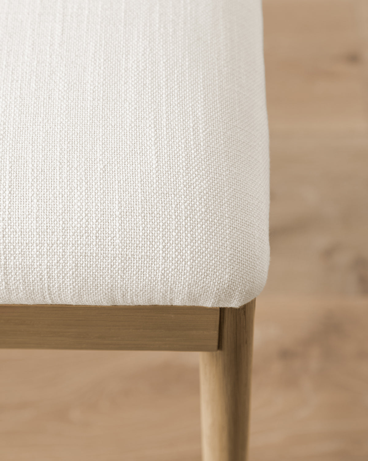 A close-up of the Molly Dining Chair shows its light beige, textured upholstery and solid oak frame with wooden legs, set on a light wood floor. The image focuses on the corner to highlight the detailed fabric and craftsmanship by McGee & Co.