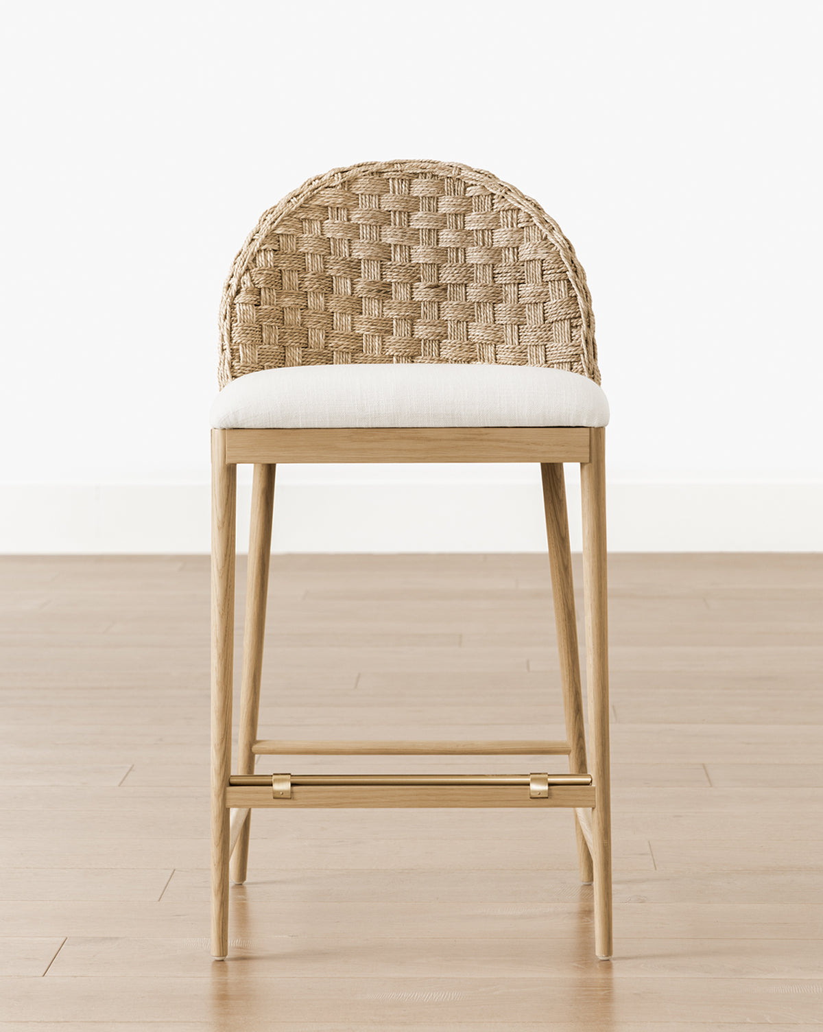 Front view of the Molly Stool, showcasing a woven rattan backrest, light cushioned seat, and solid oak legs, placed on a light wooden floor with a plain white wall in the background, by McGee & Co.