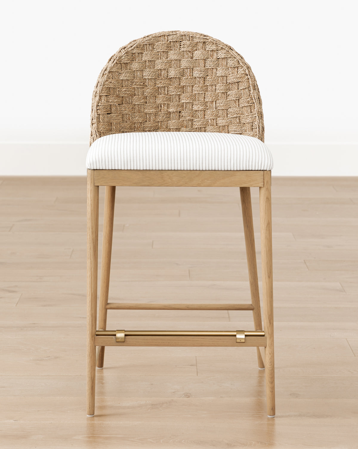 The Makerspalm Molly Stool features a woven rattan curved backrest, solid oak frame, gold-toned footrest, and a white striped cushioned seat, shown on a light wood floor against a white wall—McGee & Co.