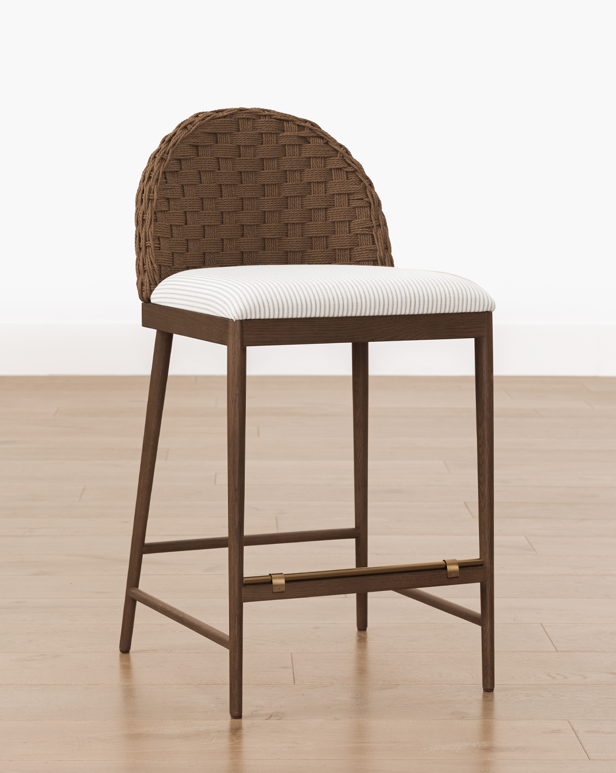 The Molly Stool features a natural finish, a woven rounded backrest, and a cushioned seat in light striped fabric, shown on a light wood floor against a plain white wall, by McGee & Co.