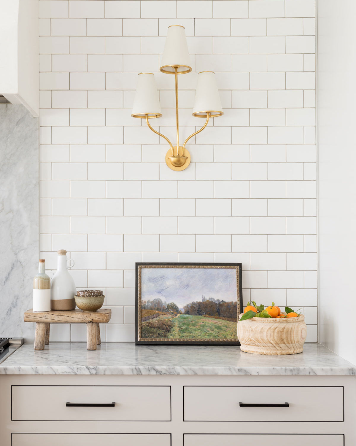 A kitchen countertop features the Impressionist Landscape artwork, a bowl of oranges, a wooden stand with bottles, and a gold wall sconce with three white shades on a white subway tile backsplash.