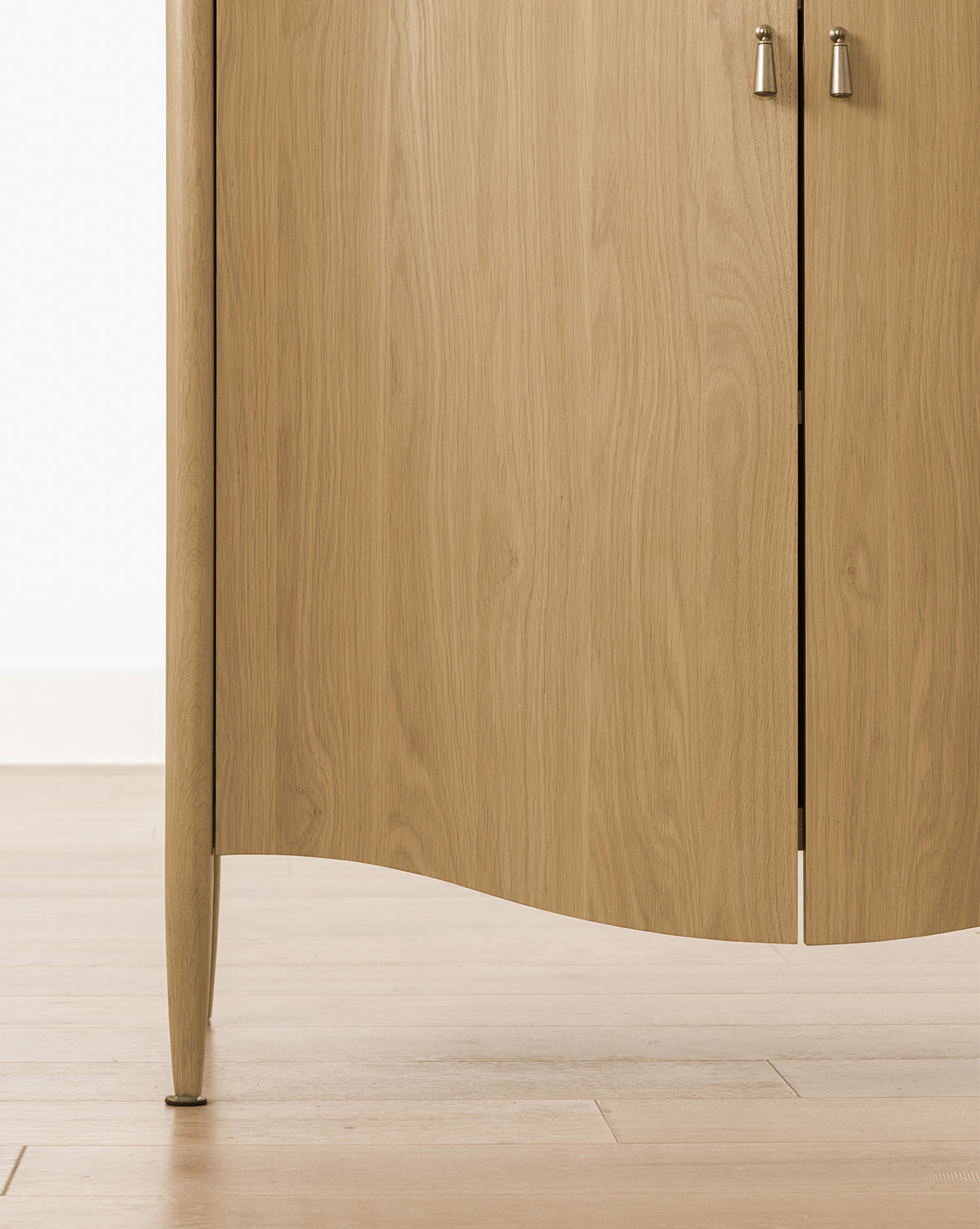 Close-up of the Moorland Cabinet with two doors and silver handles, standing on tapered legs on a light wooden floor by a white wall. Its wood grain and smooth finish make it ideal for stylish living room storage, by McGee & Co.