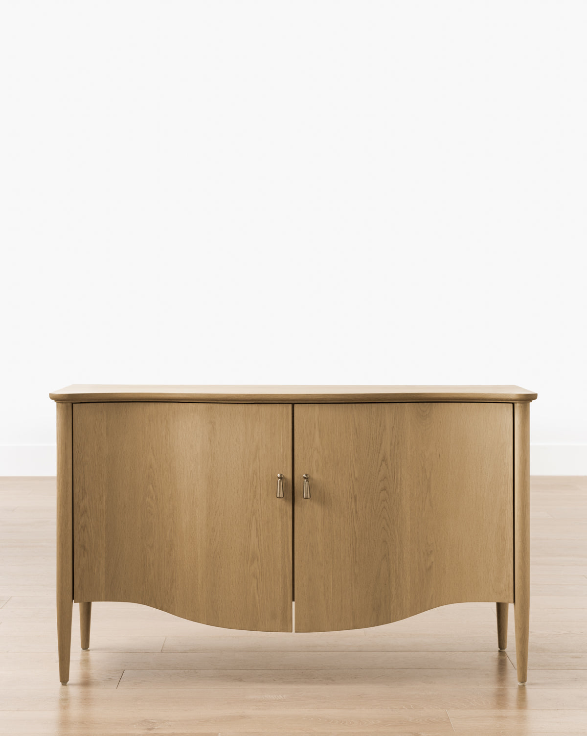The Moorland Console, crafted from white oak with two doors and silver handles, features a curved base and tapered legs, by McGee & Co. It sits on a light wood floor against a plain white wall.