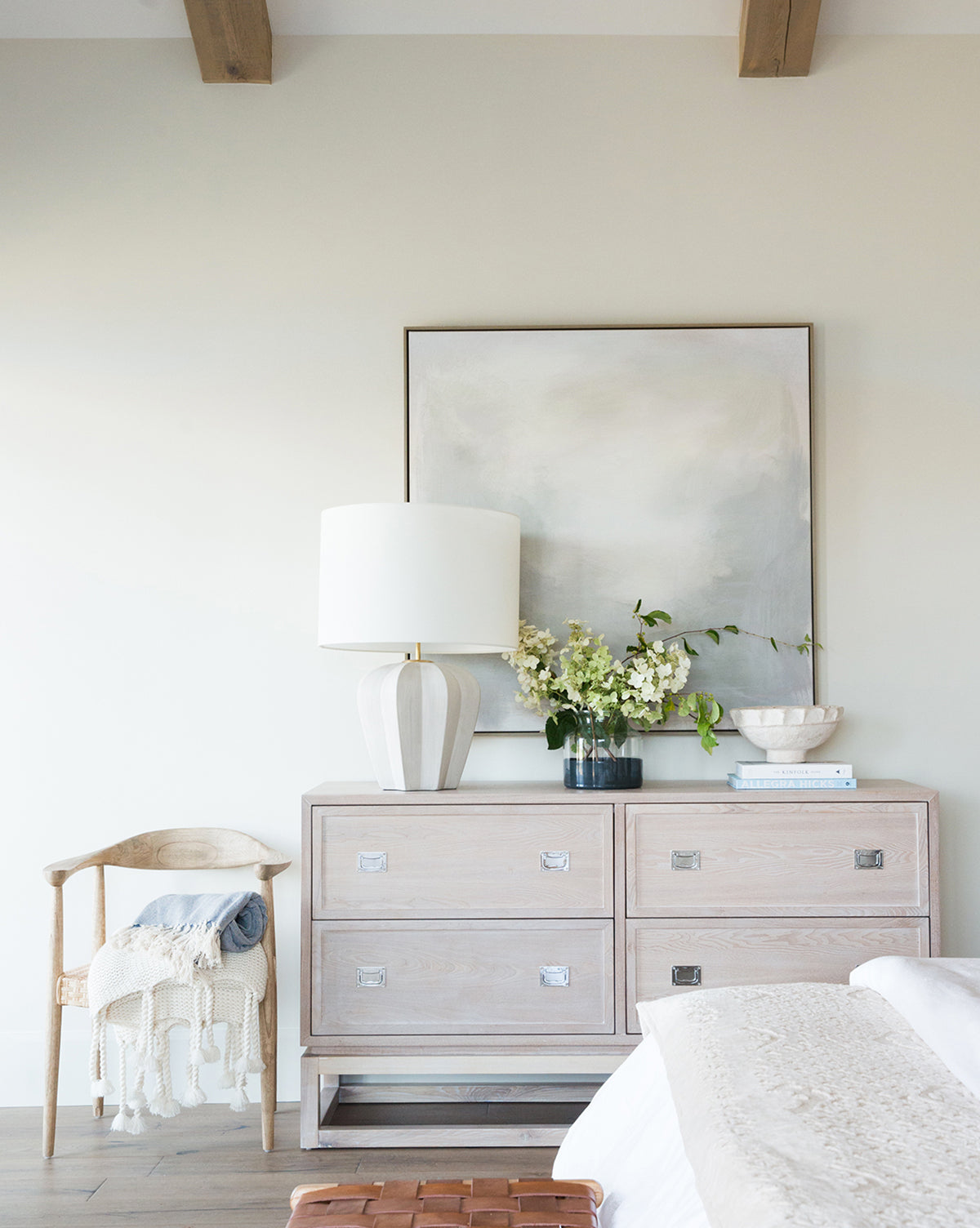 The bright bedroom features abstract Mountain Fog wall art in muted tones hanging above a light wood dresser with a white lamp, flowers, and decor, next to a wooden chair draped with a knit throw for a soothing atmosphere.