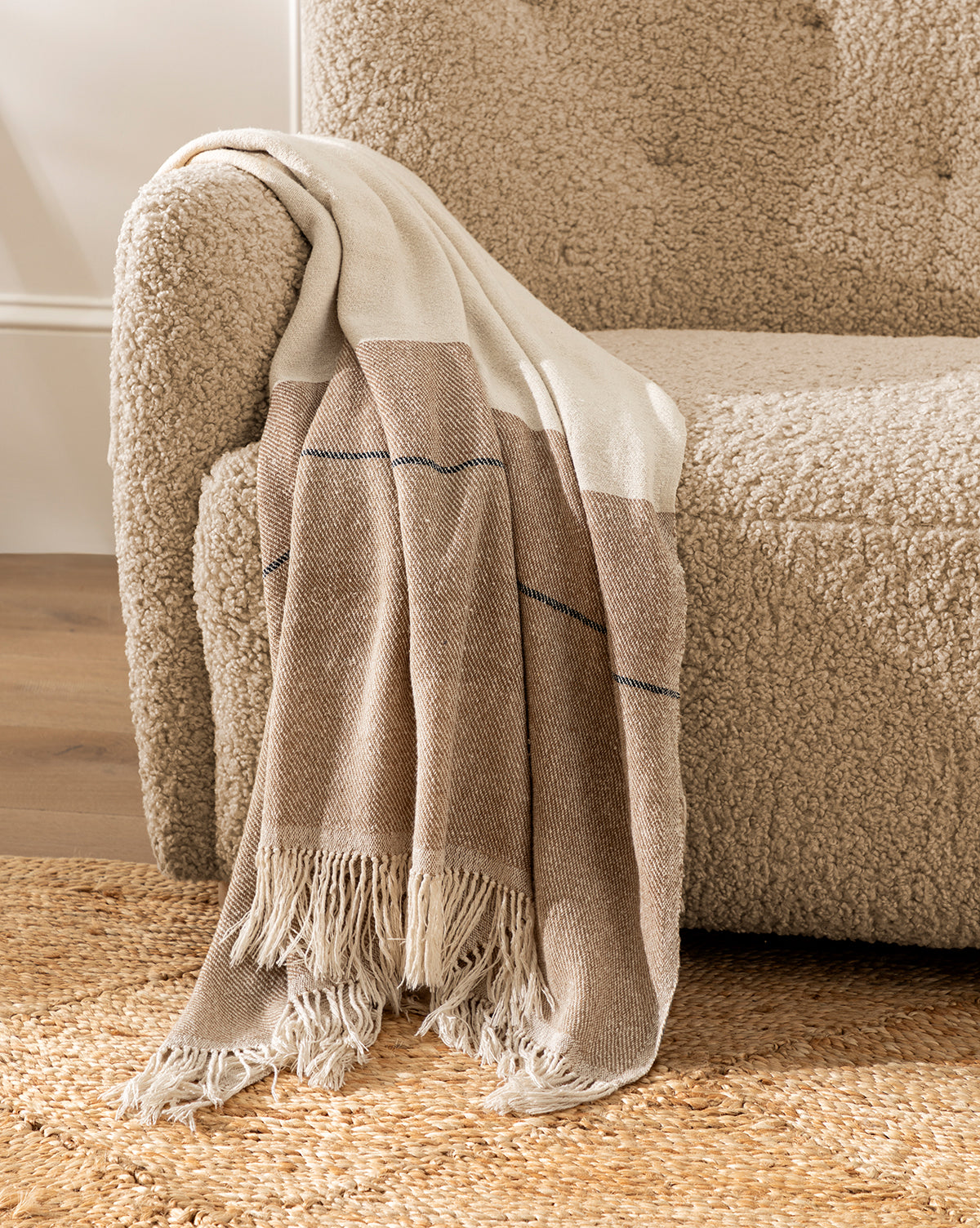 A cozy beige boucle armchair with a Murphy Silk Throw draped over the armrest sits beside a woven jute rug on a wooden floor.
