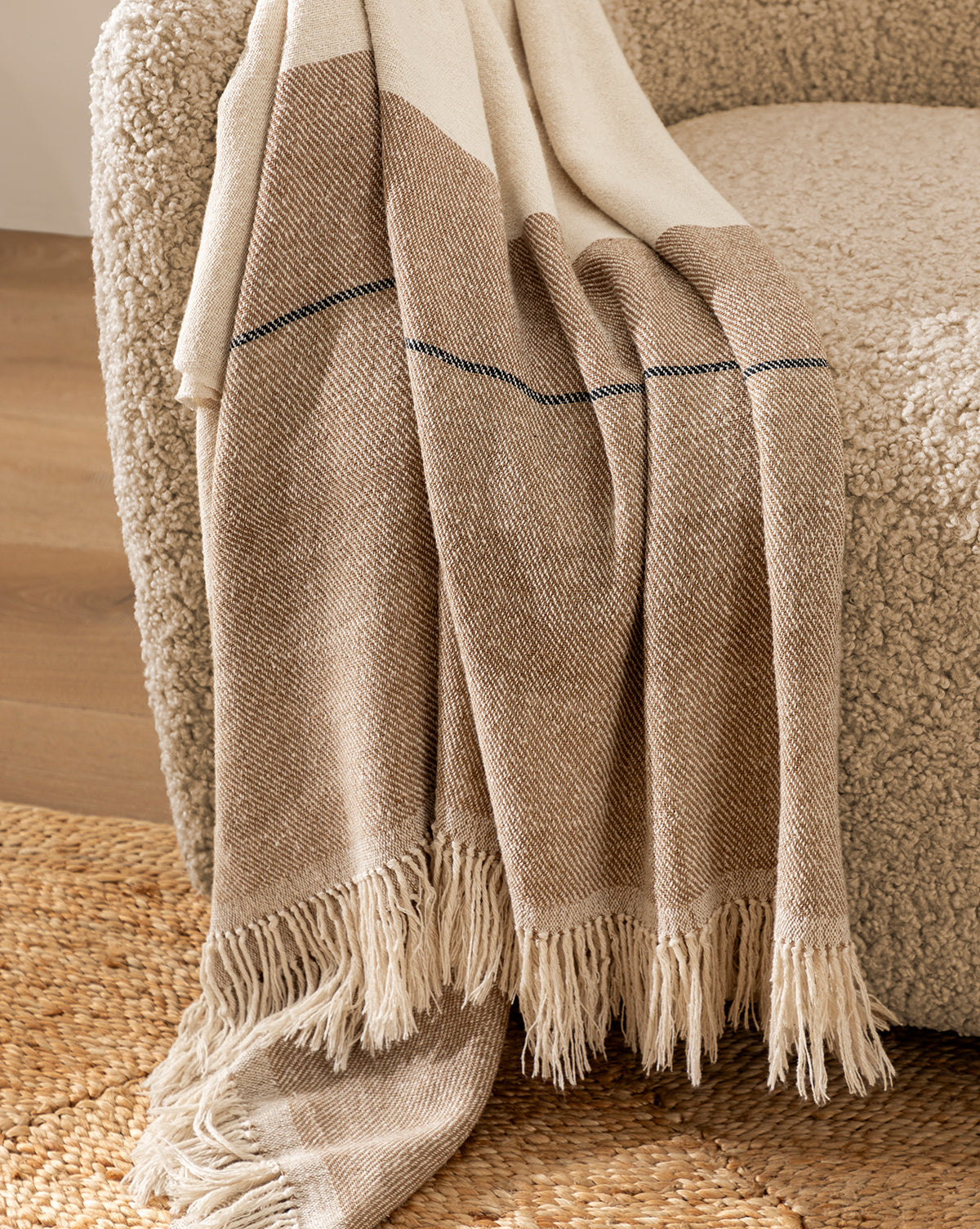 The Murphy Silk Throw, in beige and brown with fringe edges, is draped over the arm of a textured light brown chair on a woven rug above a wooden floor.