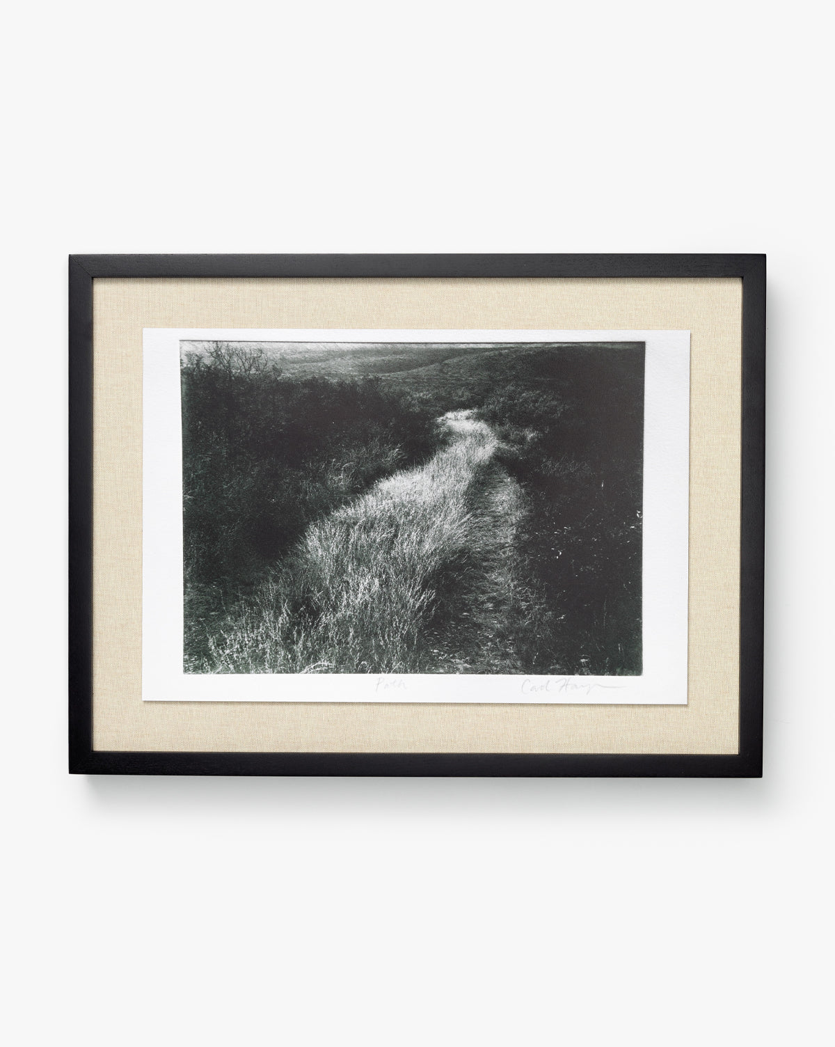 Muted Path Green by Carol Haymon is a black and white photo of a narrow grassy path through dense bushes, offered as a giclee print with a beige mat and black frame, displayed on a plain white background.