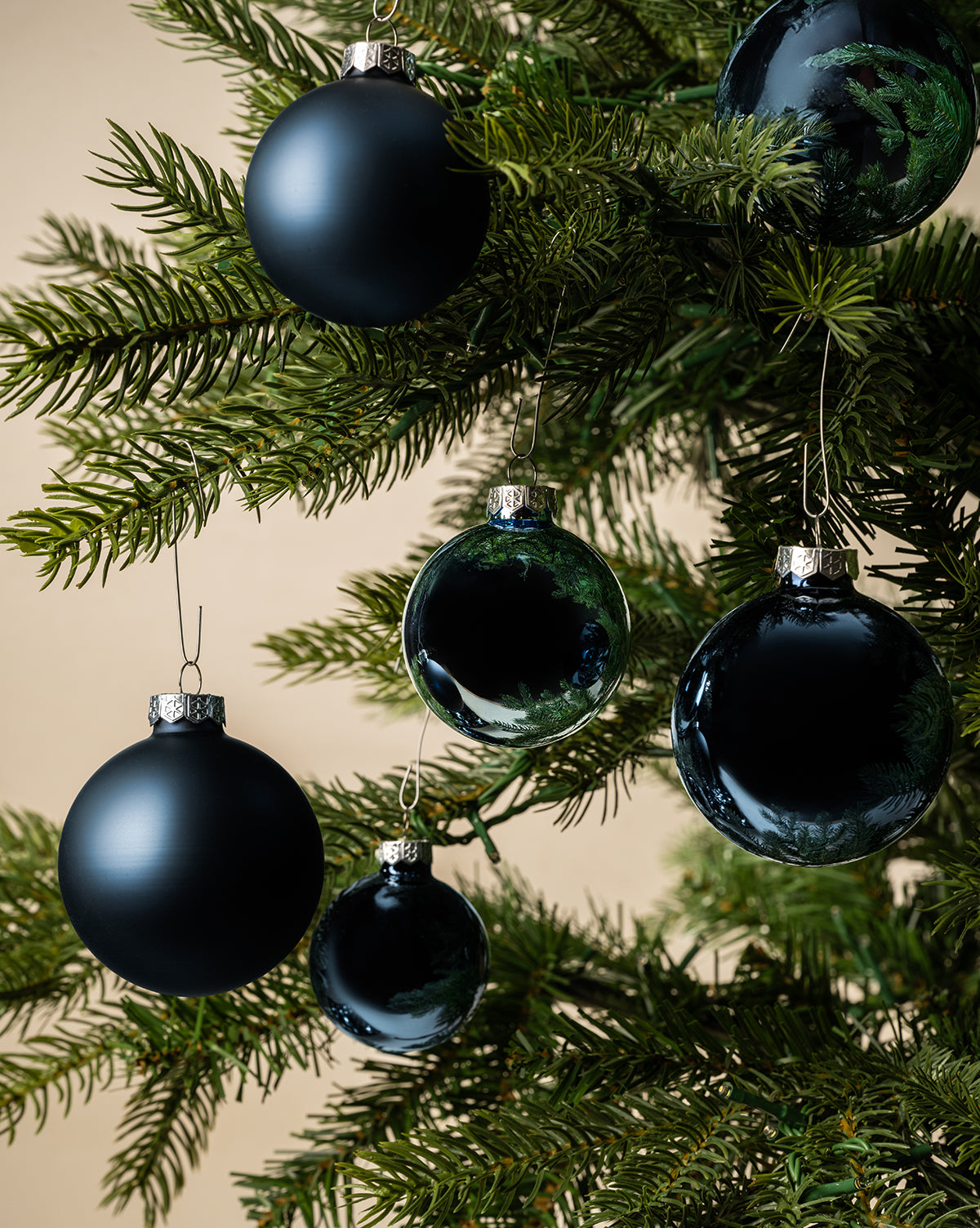 Close-up of a green Christmas tree branch decorated with Navy Glass Baubles (Set of 21 Assorted)—a mix of matte and glossy ornaments—set against a neutral beige background, by McGee & Co.