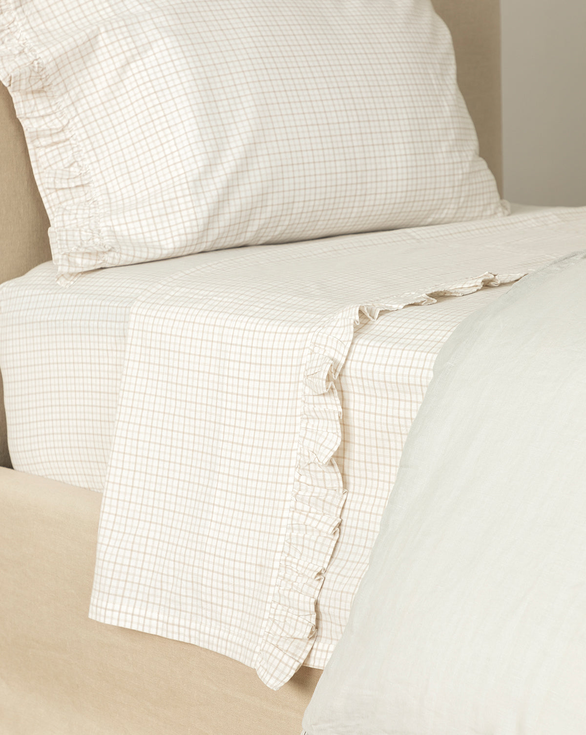 A close-up of a neatly made bed features the Nell Ruffled Twin Kids Sheet Set, with beige and white plaid cotton bedding. The pillowcase and ruffled twin sheets have delicate edges, while a light duvet completes this cozy kids set.