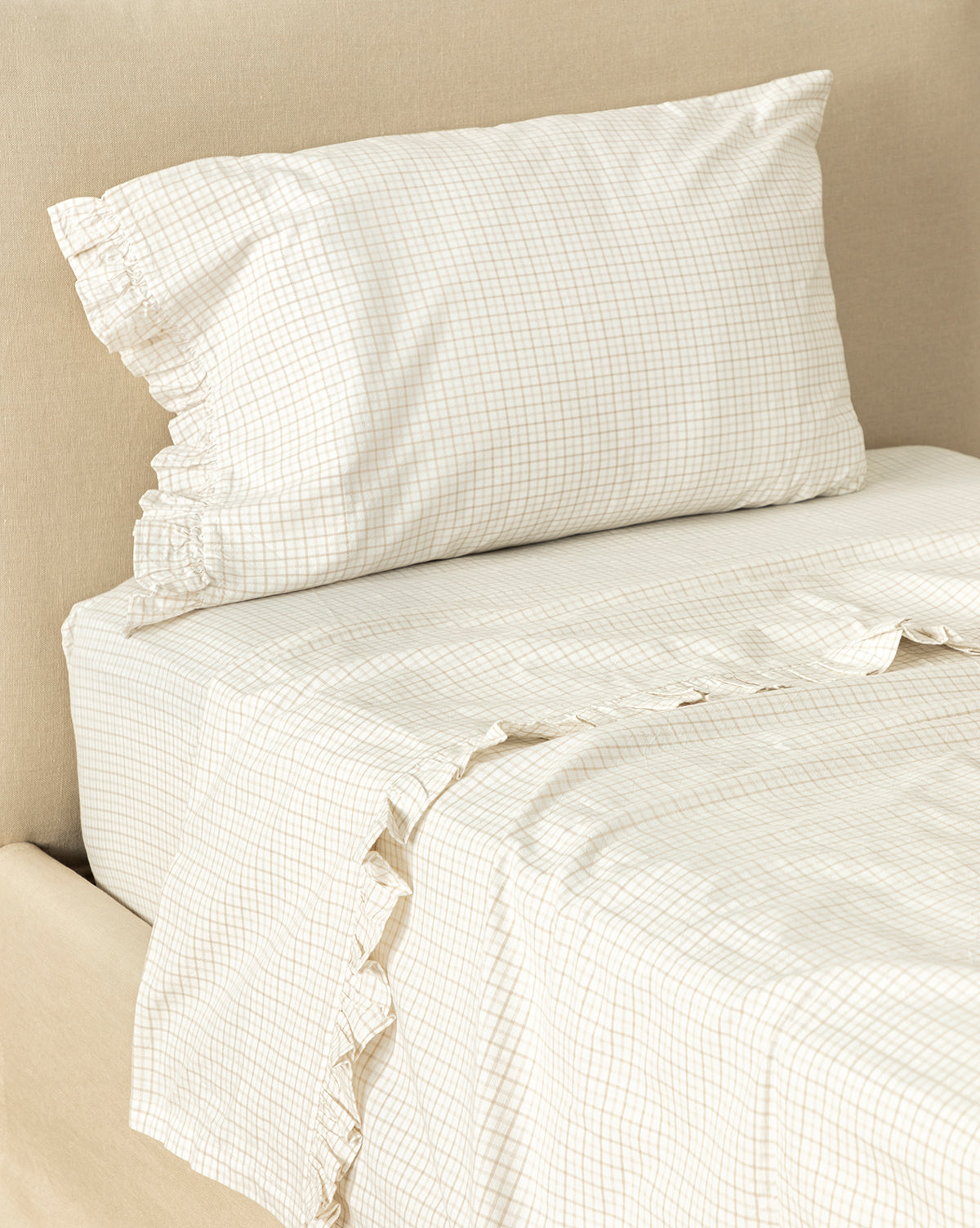 A neatly made bed features the Nell Ruffled Twin Kids Sheet Set in beige and white checks, with a ruffled pillowcase, set against a beige upholstered headboard for cozy cotton bedding.