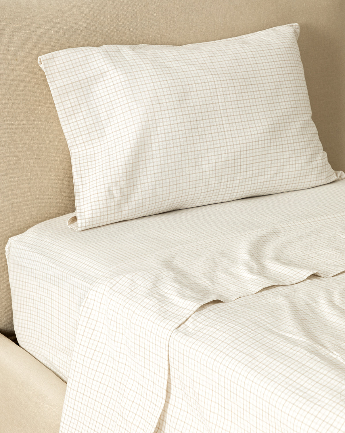 The Nell Twin Kids Sheet Set features light beige grid-patterned cotton sheets and a matching pillowcase, creating a cozy, stylish look against a light tan upholstered headboard—perfect for any kids twin bed.