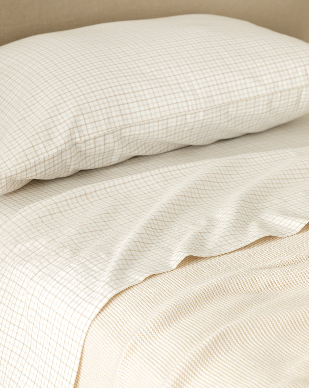 Close-up of a neatly made bed with the Nell Twin Kids Sheet Set, featuring a beige and white checkered pattern and matching pillowcase, plus a textured cream blanket underneath. The children’s bedding offers a simple, minimalist design.
