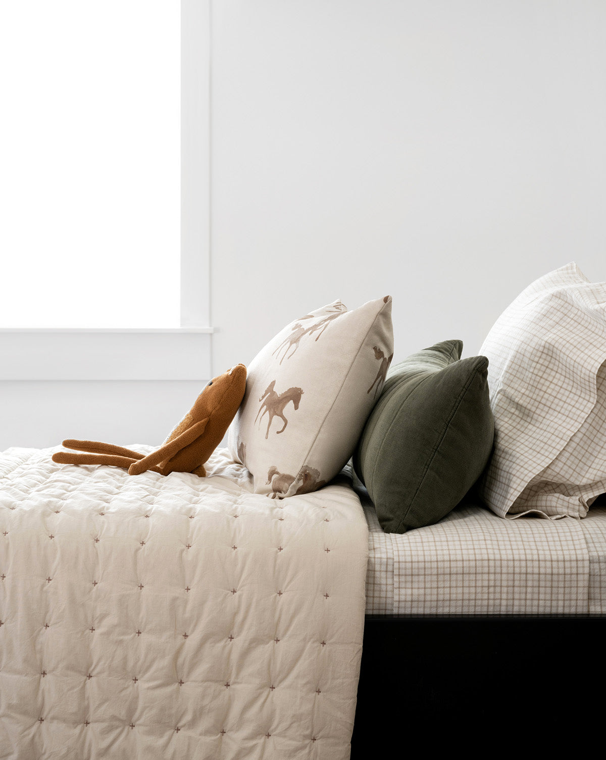A bed with a beige quilt, the Nell Twin Kids Sheet Set in cotton plaid, two pillows—one with a horse design and one green—and a soft brown rabbit toy on top, all by a window against white walls.