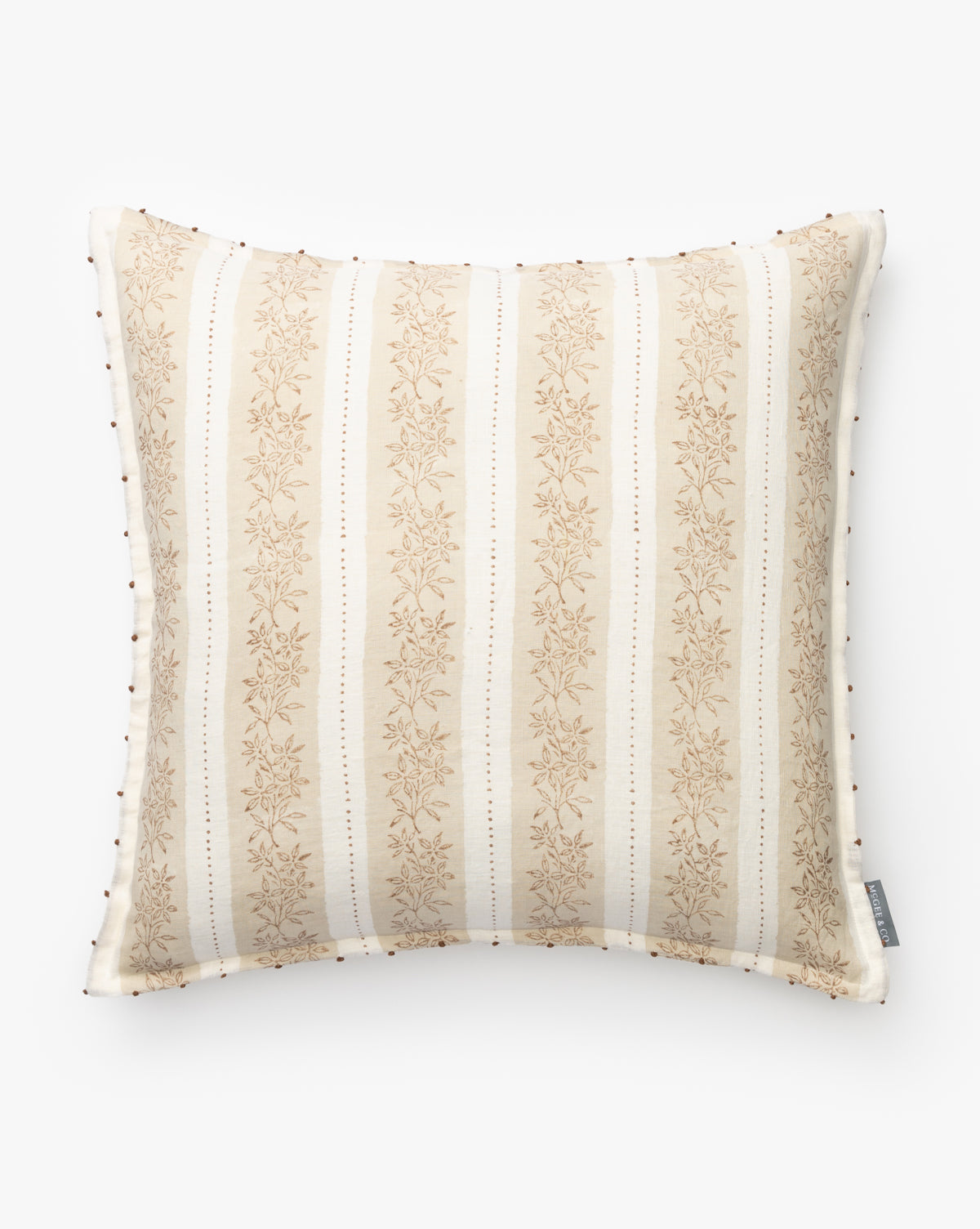 The Nettles Pillow Cover is a square linen cover with cream and beige vertical stripes, a subtle floral pattern, small decorative beads on the edges, and a fabric tag on the right side. Shown against a plain white background by McGee & Co.