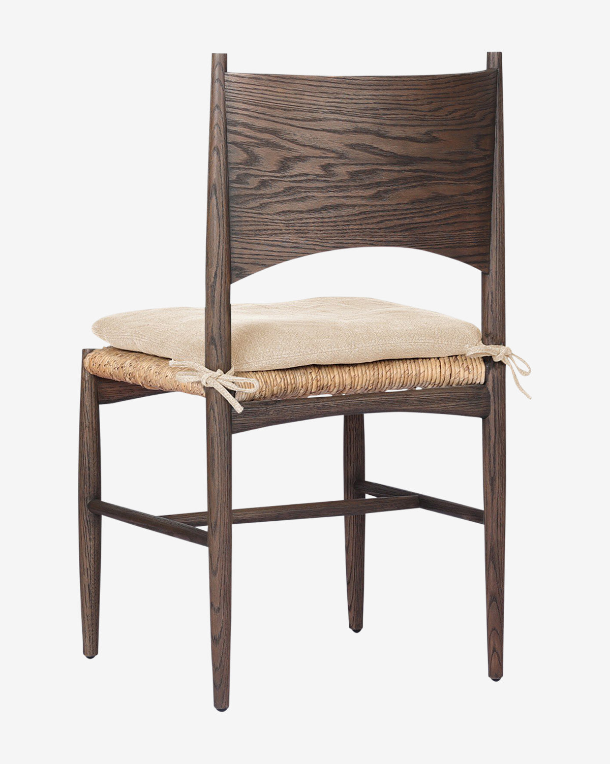 The Nixie Dining Chair features a dark solid oak frame, a woven banana leaf seat, and a light, high-performance fabric cushion tied to the chair. Its wide curved backrest and slim tapered legs create an elegant silhouette against a white background by McGee & Co.