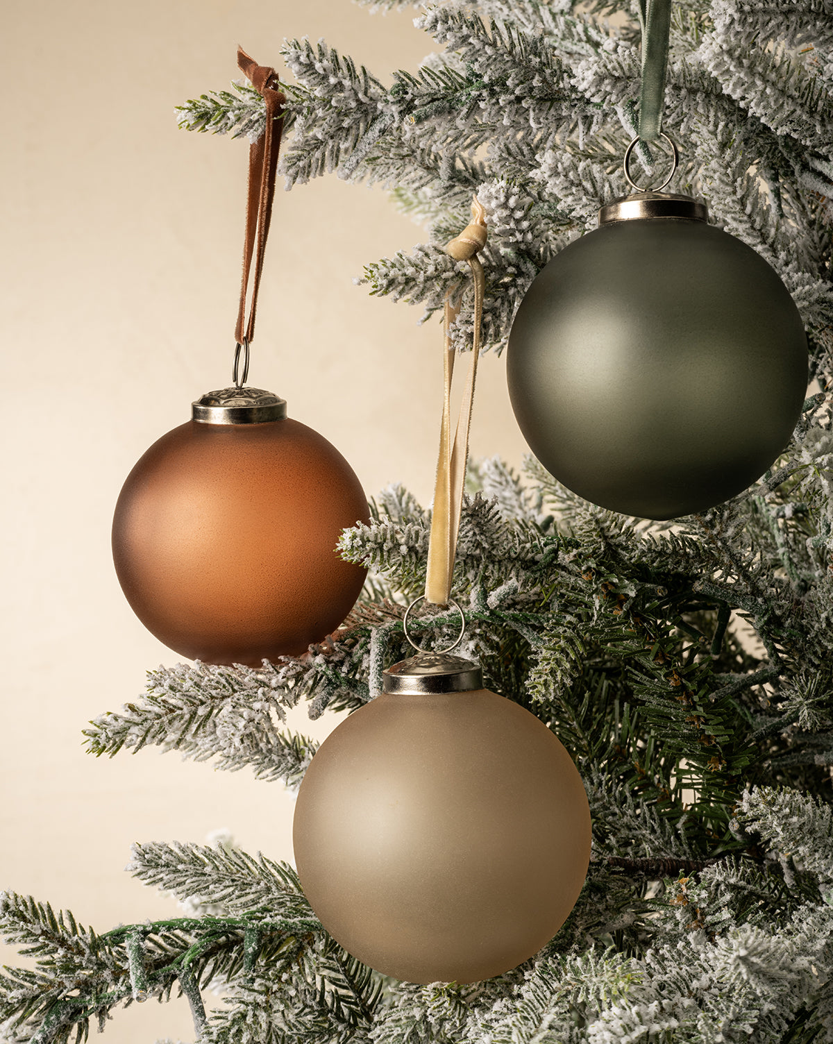 Three frosted Noel Festive Ornaments from the 9-piece set—brown, green, and gold glass—hang on a snow-dusted artificial Christmas tree against a neutral background, by McGee & Co.