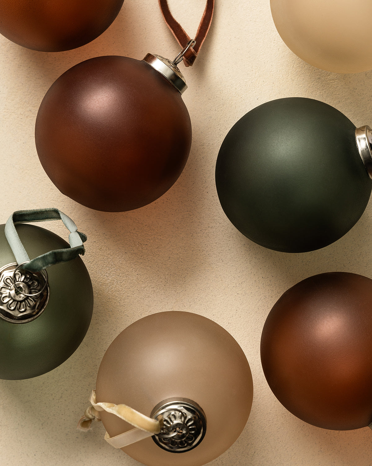 A flat lay features the Noel Festive Ornaments (Set of 9) in matte glass with earthy brown, green, and beige tones, each with decorative silver caps and assorted ribbon hangers, arranged on a neutral background for Christmas décor, by McGee & Co.
