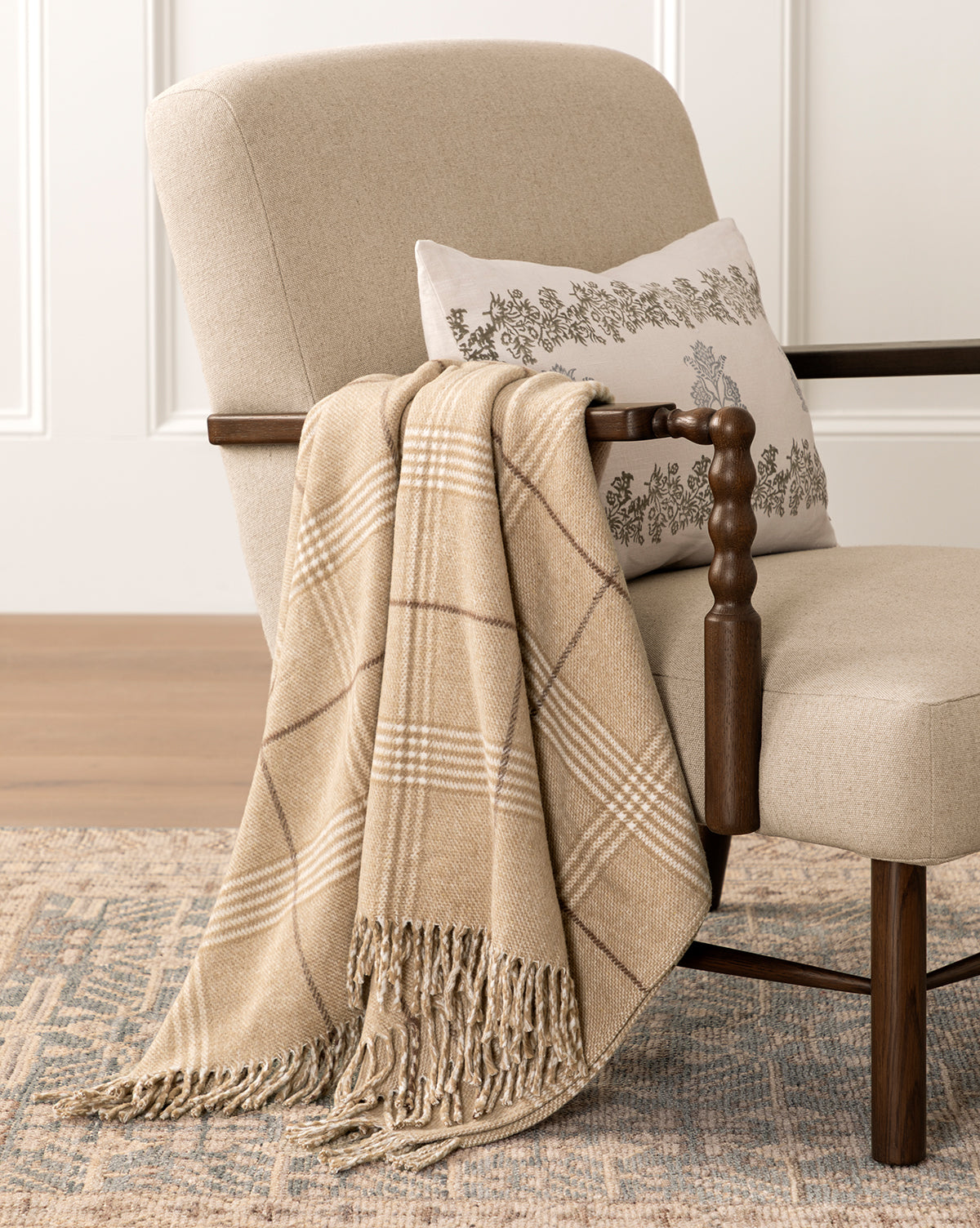 A beige armchair with wooden armrests features a cream floral pillow and the Nottingham Throw, a tan plaid blanket, bringing cozy home decor to the softly lit room. The chair is set on a patterned rug, by McGee & Co.