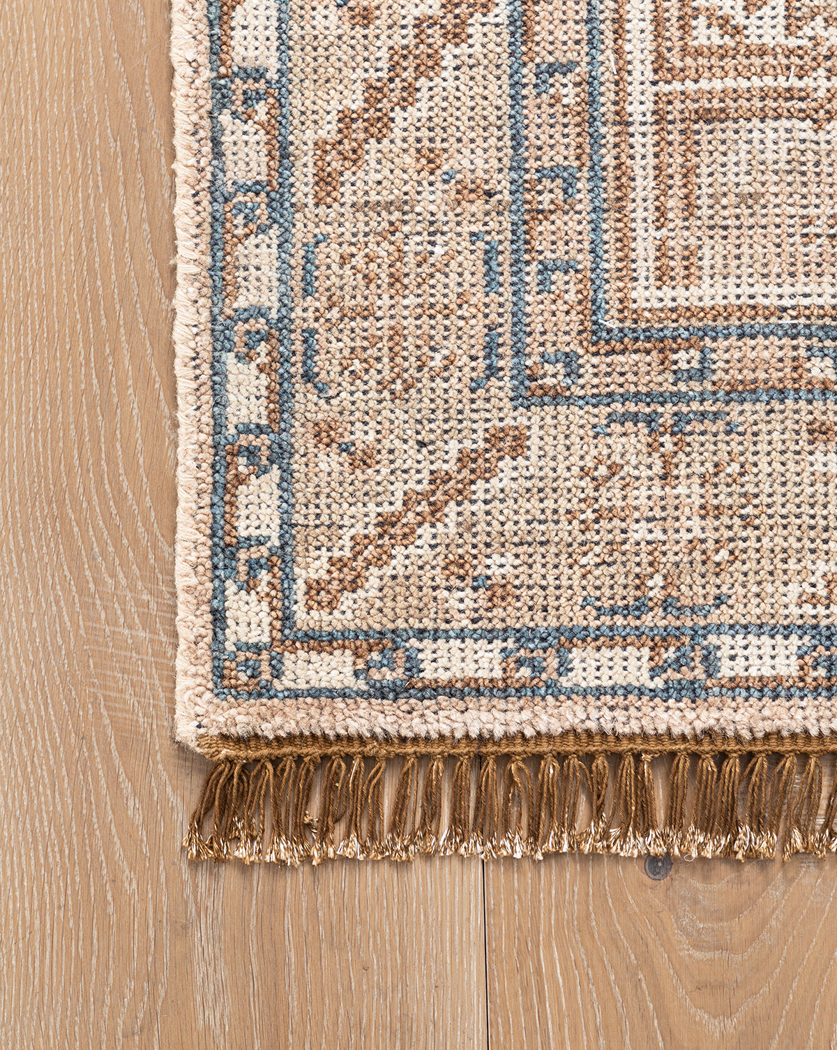 Close-up of the Nunez Hand-Knotted Wool Rug shows its beige, brown, and blue geometric pattern, tasseled fringe, and classic border design, displayed on a light wooden floor, by McGee & Co.