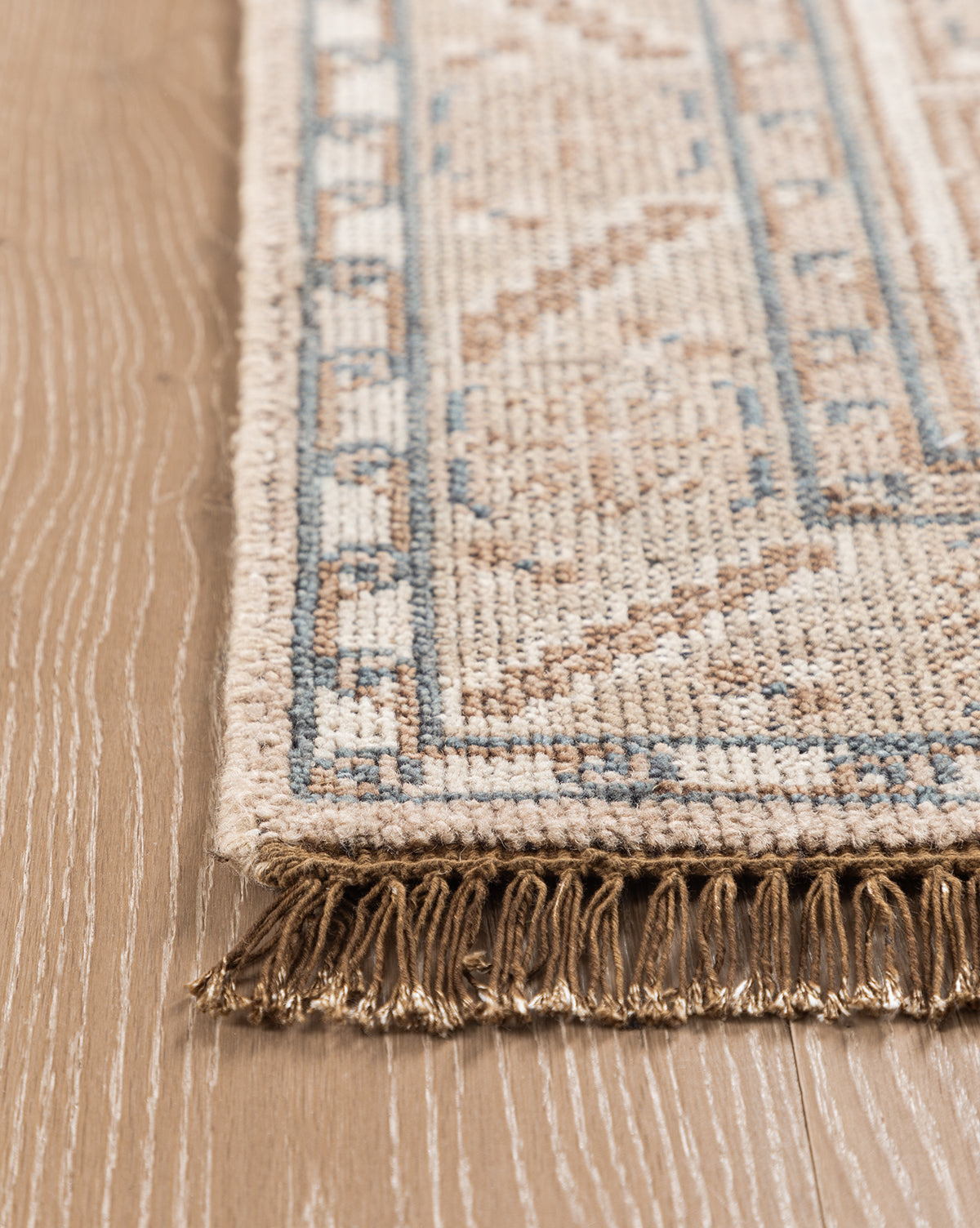A close-up of the Nunez Hand-Knotted Wool Rug shows its beige and blue geometric pattern, fringed edges, and classic border design, beautifully placed on a light wood floor, by McGee & Co.