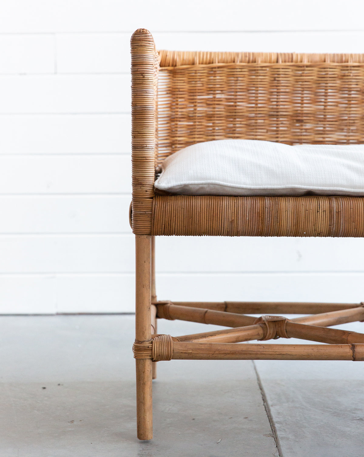 A close-up side view of the Odelia Bench, featuring a rattan frame and white cushion on a concrete floor against a white wooden plank wall. A performance fabric pillow completes the stylish, comfortable look.