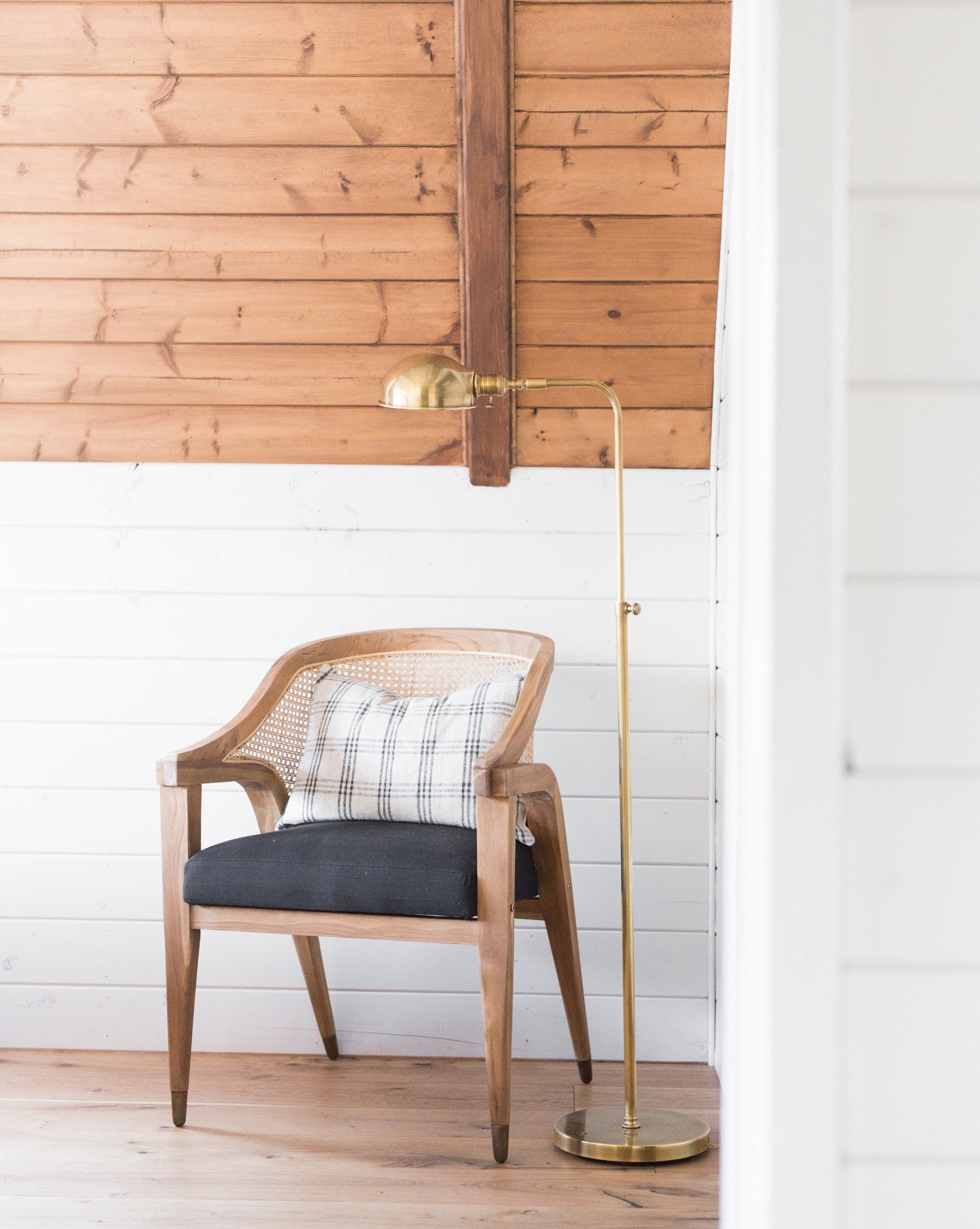 An Old Pharmacy Floor Lamp stands next to a wooden chair with a plaid cushion and dark seat in a cozy corner featuring white shiplap walls and a sloped wooden ceiling, by McGee & Co.