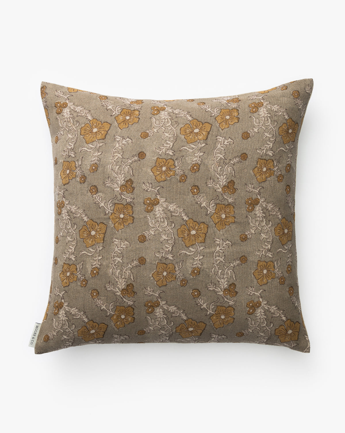 The Orchid Floral Pillow Cover features a beige background with an intricate block-print floral pattern in mustard yellow and light taupe, by McGee & Co. A small fabric tag is sewn into one side. The cover is shown on a white surface.