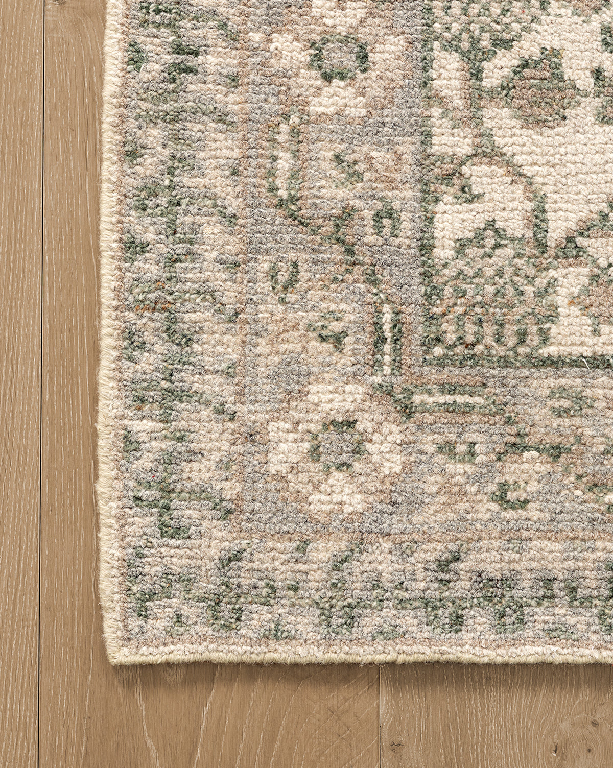 Close-up of the corner of the Orsino Hand-Knotted Rug in green, beige, and cream on a light wood floor, showcasing its antique style with a decorative border featuring floral and geometric motifs, by McGee & Co.