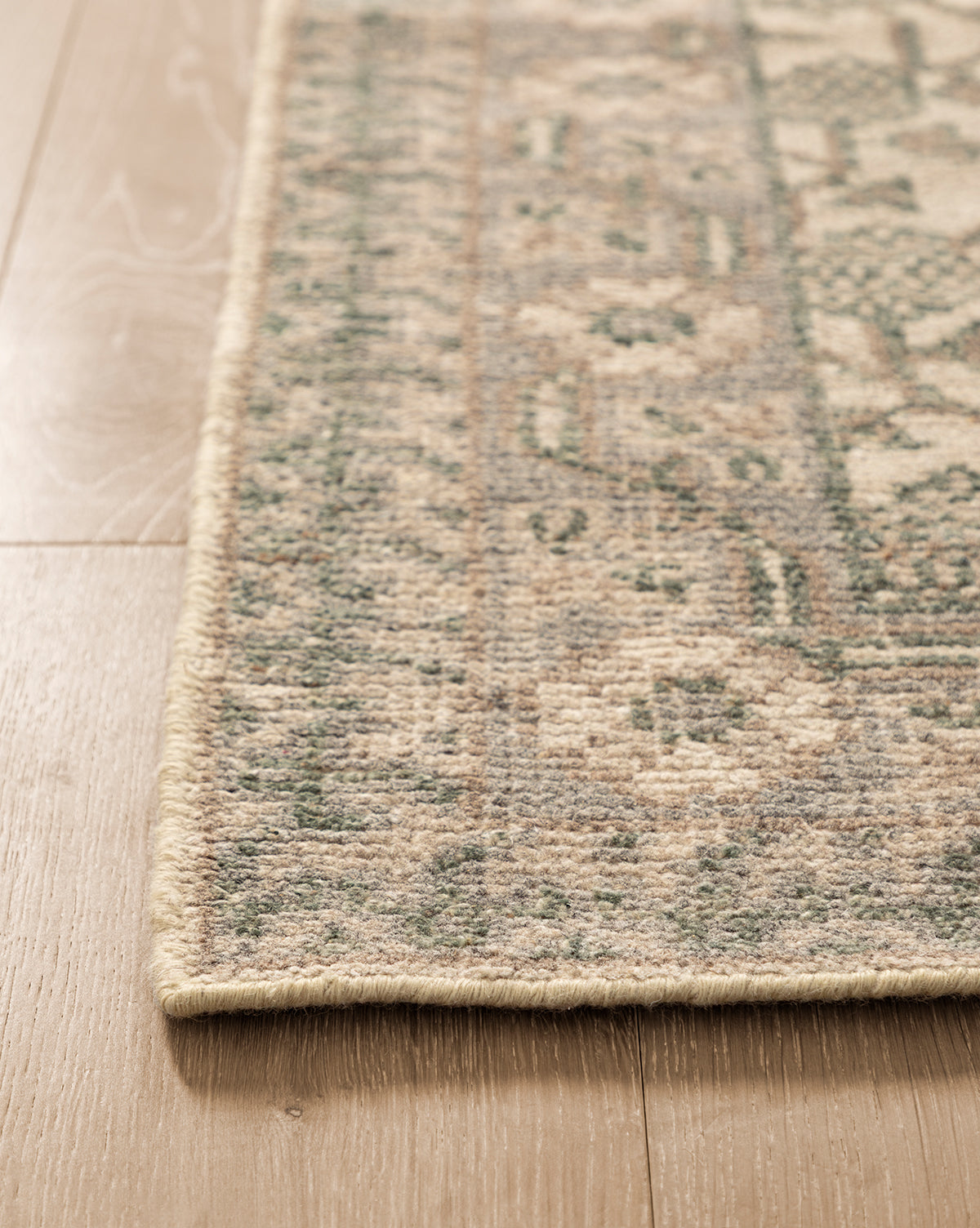 Close-up of the Orsino Hand-Knotted Rug, featuring green and beige patterns, lying flat on a light wood floor. Its antique-style design is highlighted by a neatly stitched border along the corner, by McGee & Co.
