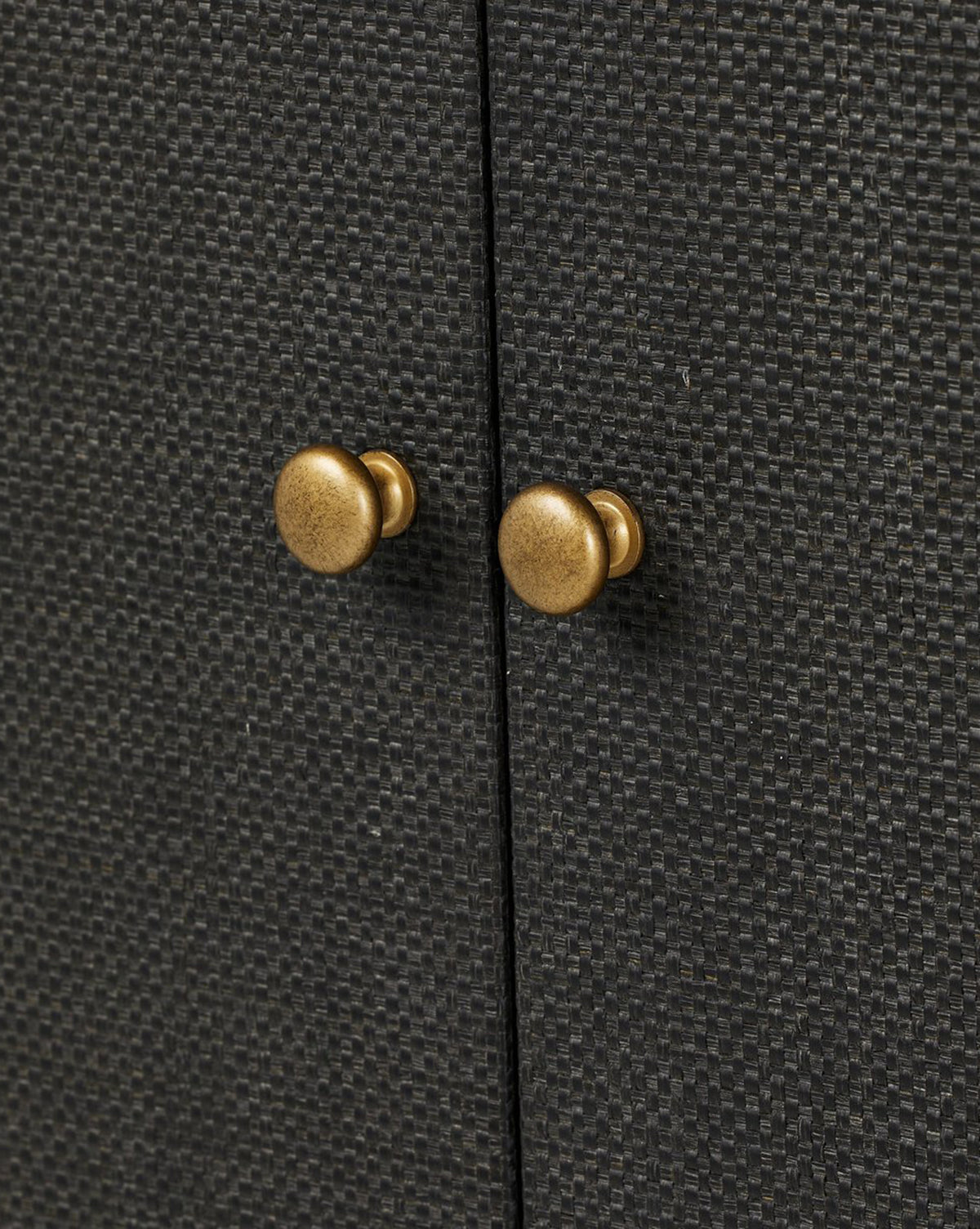 Close-up of two round, antique brass knobs on the textured black raffia surface of the Osborne Cabinet. The cabinet doors meet in the center between the hardware, by McGee & Co.