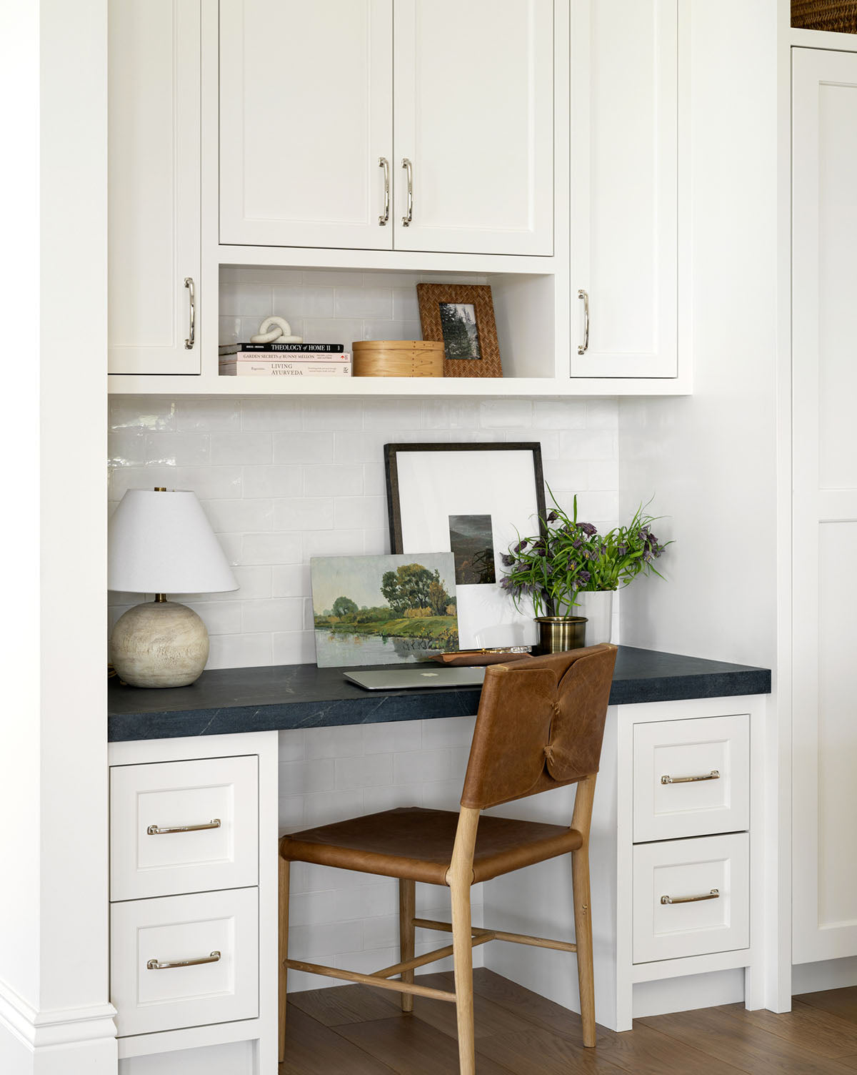 A compact workspace features white cabinetry, a black countertop, the Danson Dining Chair with a leather seat, a table lamp, framed art, books, and a potted plant for a cozy, minimalist look by McGee & Co.