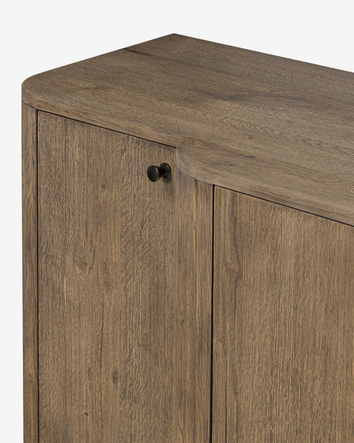 Close-up of the Oswin Sideboard in weathered oak with a natural brown finish, by McGee & Co. Shown are part of the top and two doors, one with a small round black knob. The cabinet has a modern break-front design and rounded edges.