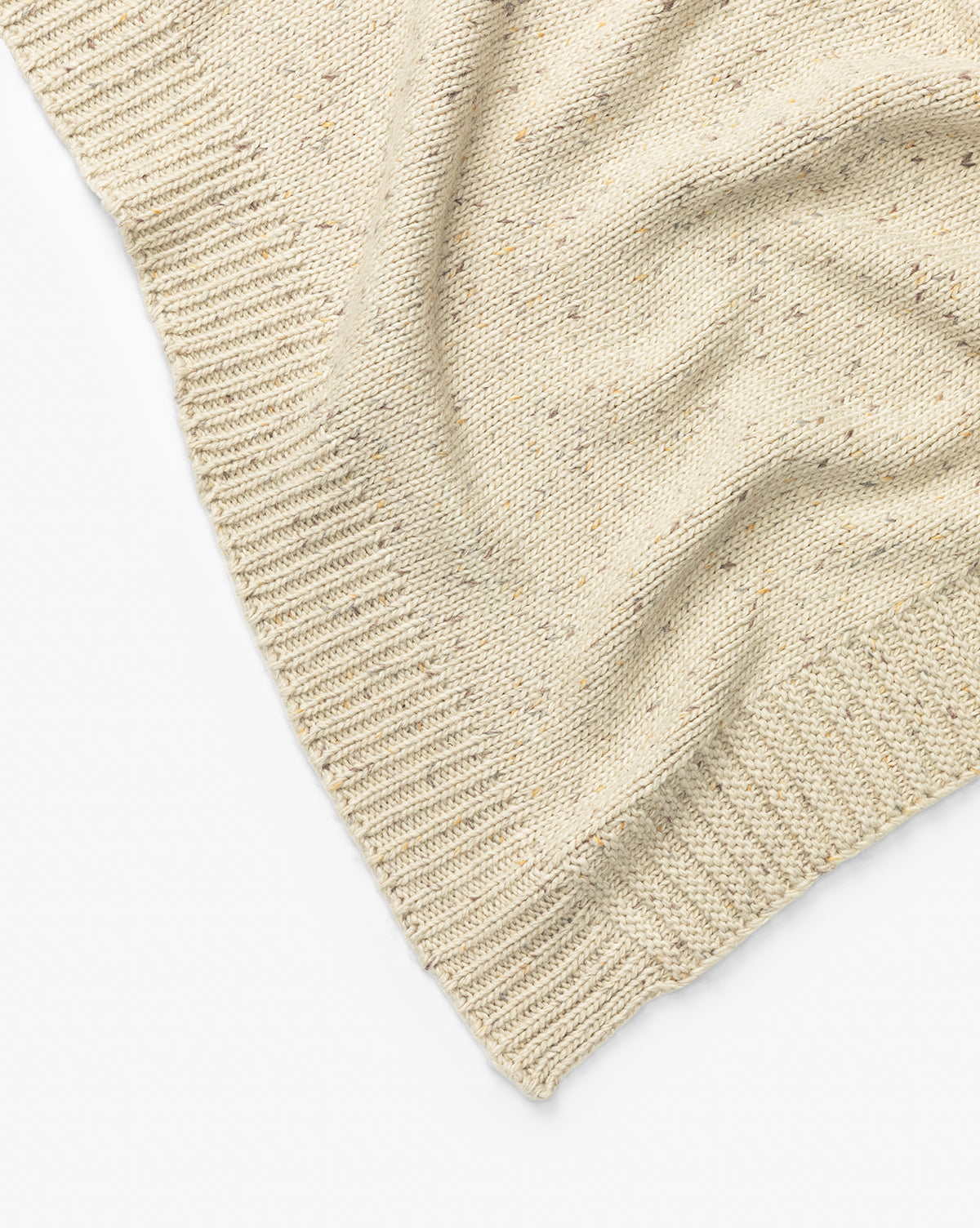 A close-up of the Makerspalm Otis Knit Throw shows its beige wool blend with ribbed edges, a subtly wavy texture, and small grey-speckled flecks throughout. McGee & Co.