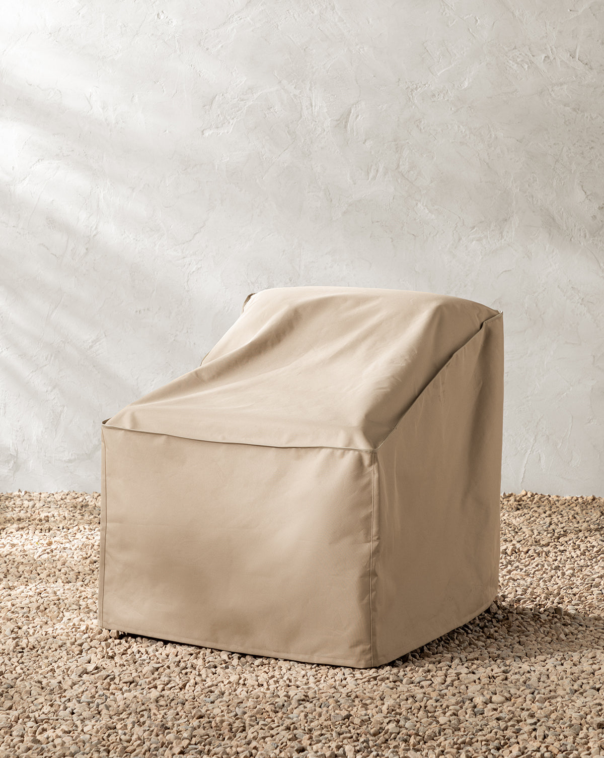 A beige, boxy bean bag chair with a backrest sits on a textured pebble floor against a light wall, protected by the Outdoor Lounge Chair Cover, which is water-resistant and designed for outdoor use, by McGee & Co.