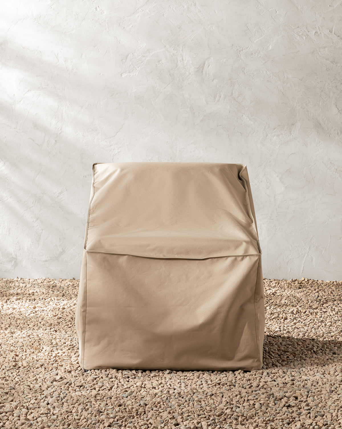 A beige, boxy chair with a leather-like texture sits on pebbled flooring against a light wall, shielded from the elements by the water-resistant Outdoor Lounge Chair Cover, by McGee & Co.