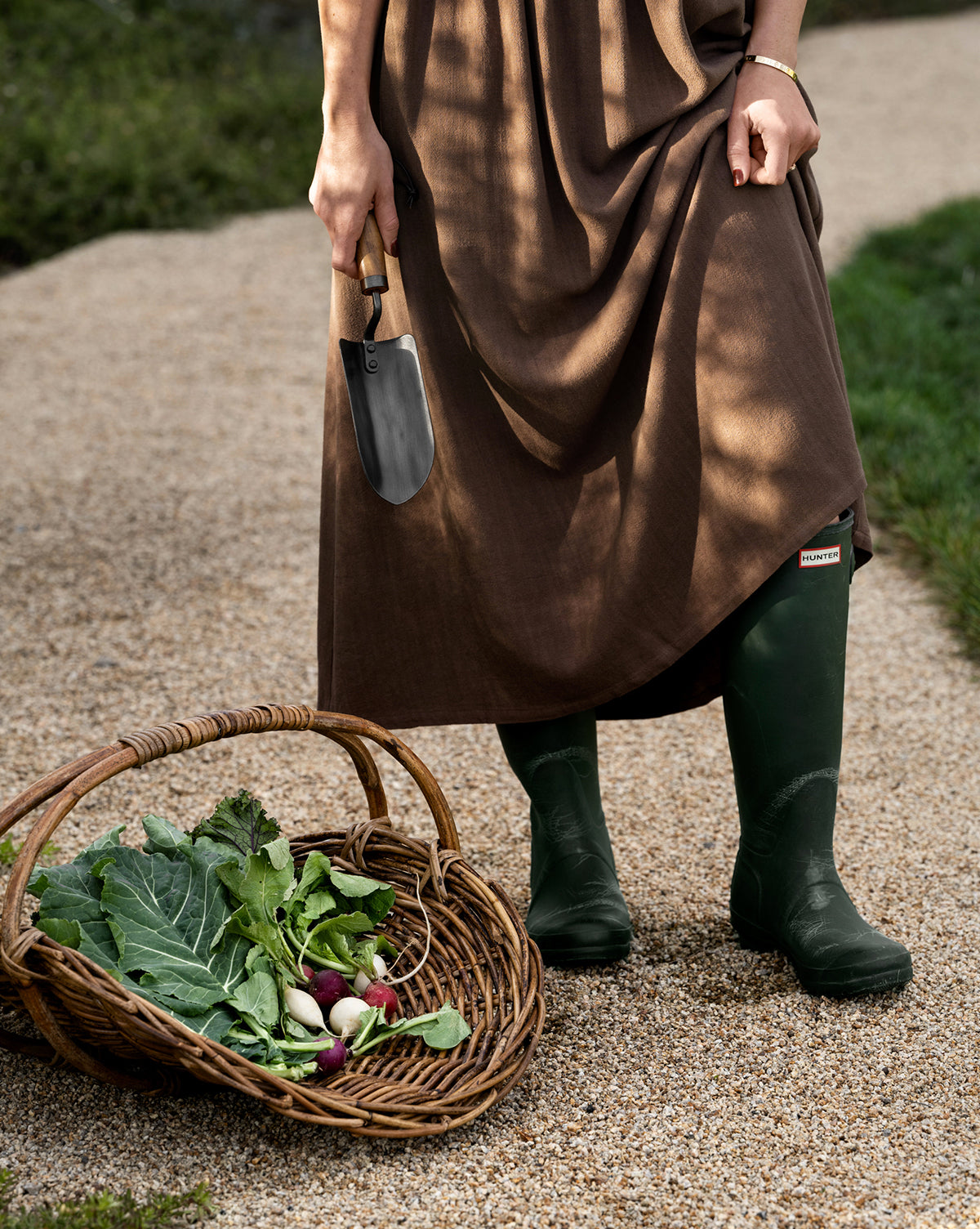 Wearing a brown dress and green boots, a person stands on a gravel path holding a trowel next to a Rattan Gardening Tray filled with fresh vegetables, creating an organic vibe.