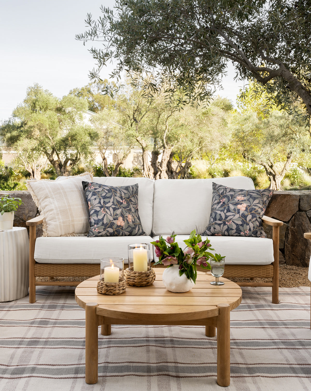A cozy outdoor patio features a wooden sofa with white cushions and floral pillows, plus a round wooden coffee table accented by the Woven Hurricane candle display, all set amid trees and greenery for a natural touch.