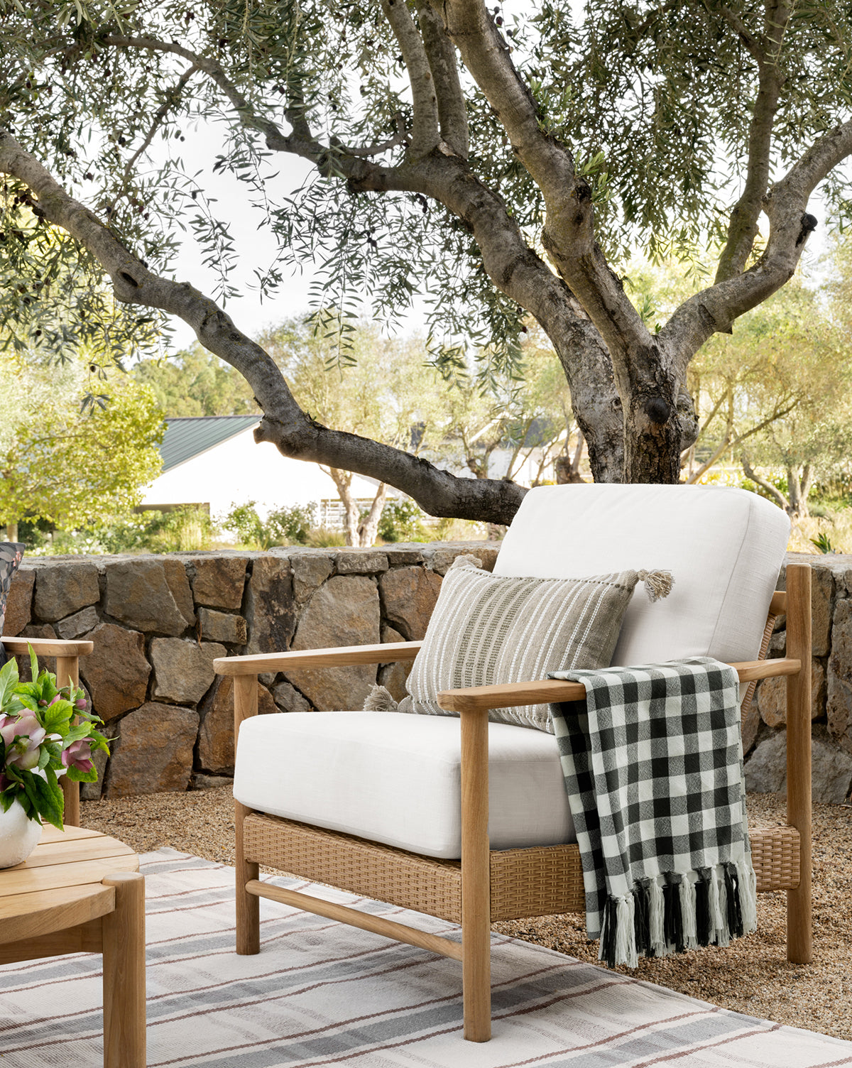 The Simeon Outdoor Lounge Chair, featuring upholstered cushions, a striped pillow, and a black-and-white checkered throw, sits on the patio by a stone wall and large tree amidst lush plants and greenery, by McGee & Co.