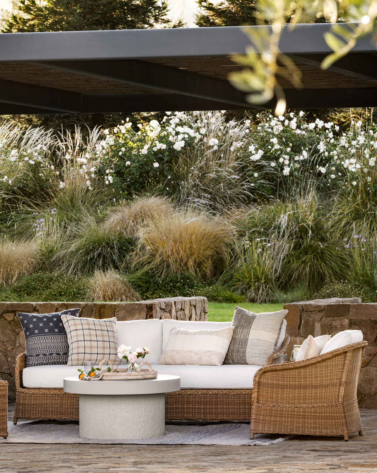A cozy outdoor seating area showcases the Haviland Outdoor Sofa 88 with neutral pillows, a round white coffee table with flowers, and lush greenery plus white flowering plants in the background by McGee & Co.