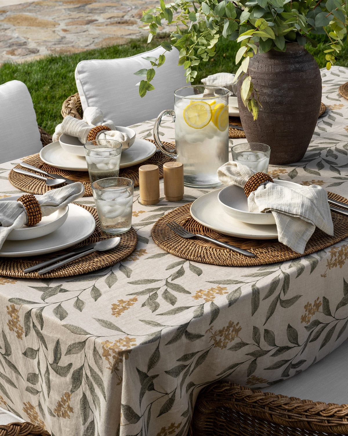 An outdoor table set for four features Davin Placemats, white plates, beige napkins in wooden rings, glasses of water, a pitcher of lemon water, salt and pepper shakers, and a large vase with greenery on a floral tablecloth, by McGee & Co.