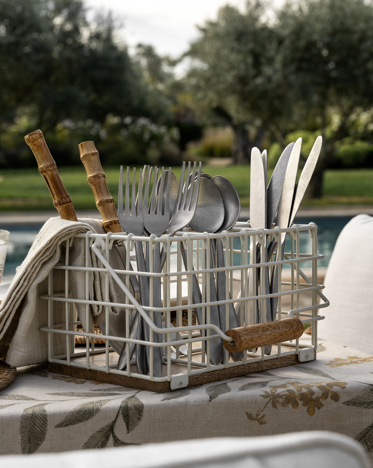 The Lilia Flatware (Set of 5) is held in a metal wire caddy with wooden handles, placed on a floral tablecloth outdoors by a pool, with trees in the background.